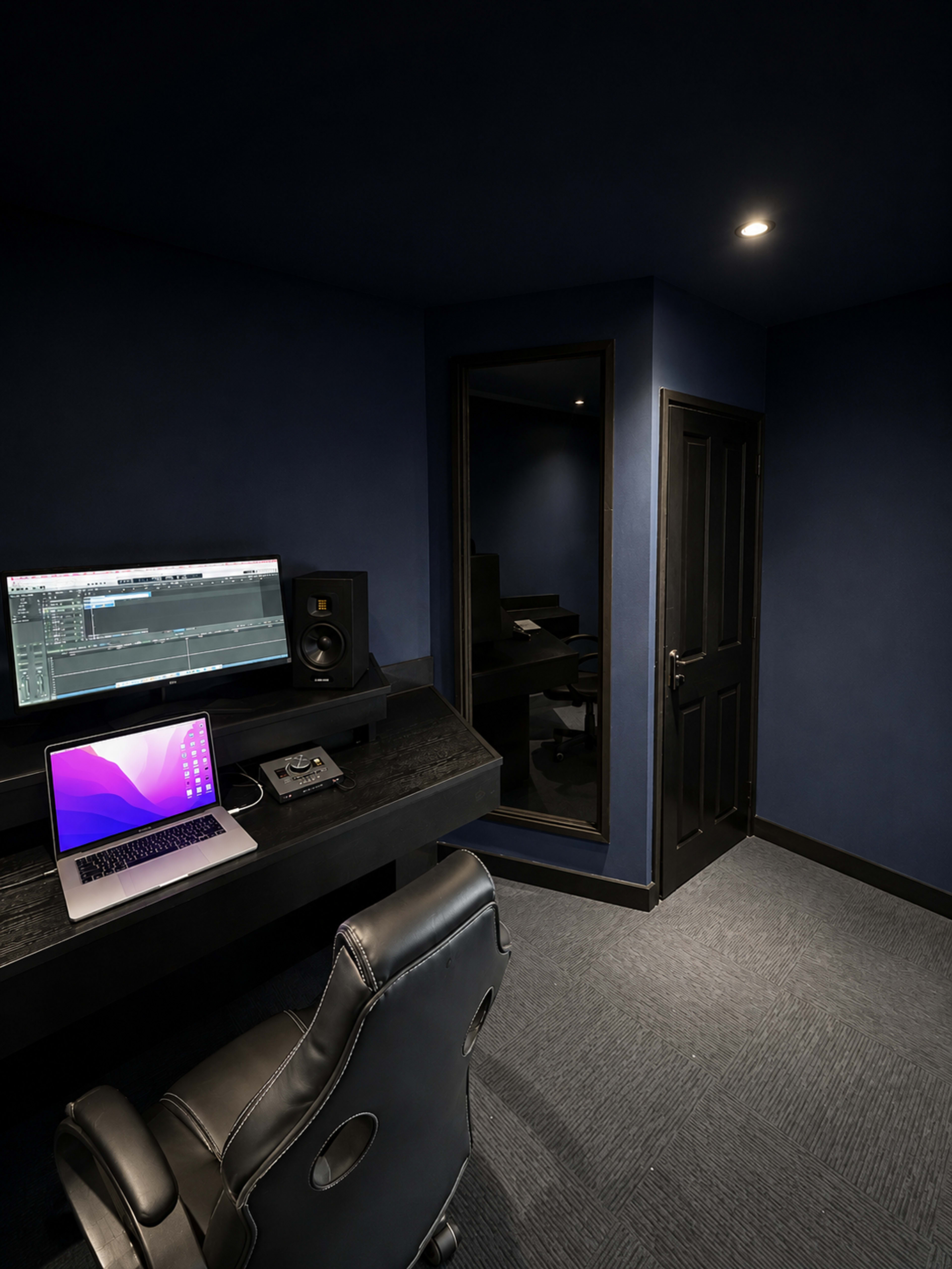 The image shows a dimly lit recording studio with a desk featuring a laptop and audio equipment, adjacent to a mirrored wall and a closed black door.