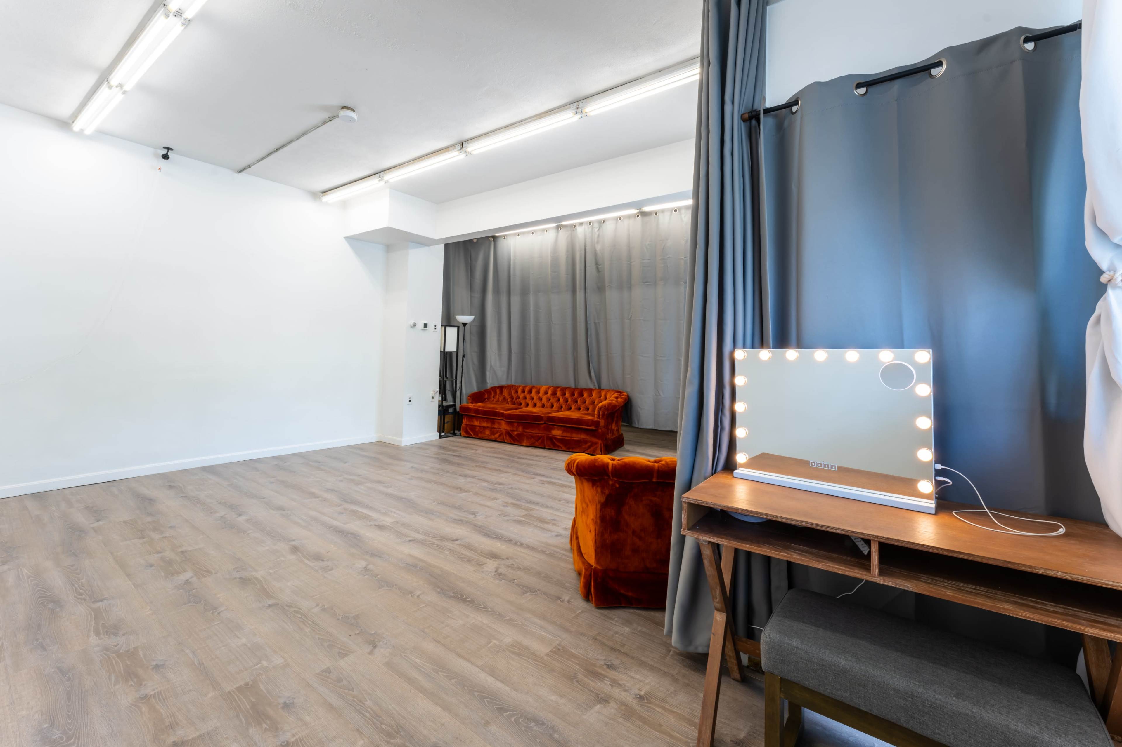 A spacious room with wooden flooring, a velvet sofa, a makeup mirror with lights, and gray curtains.