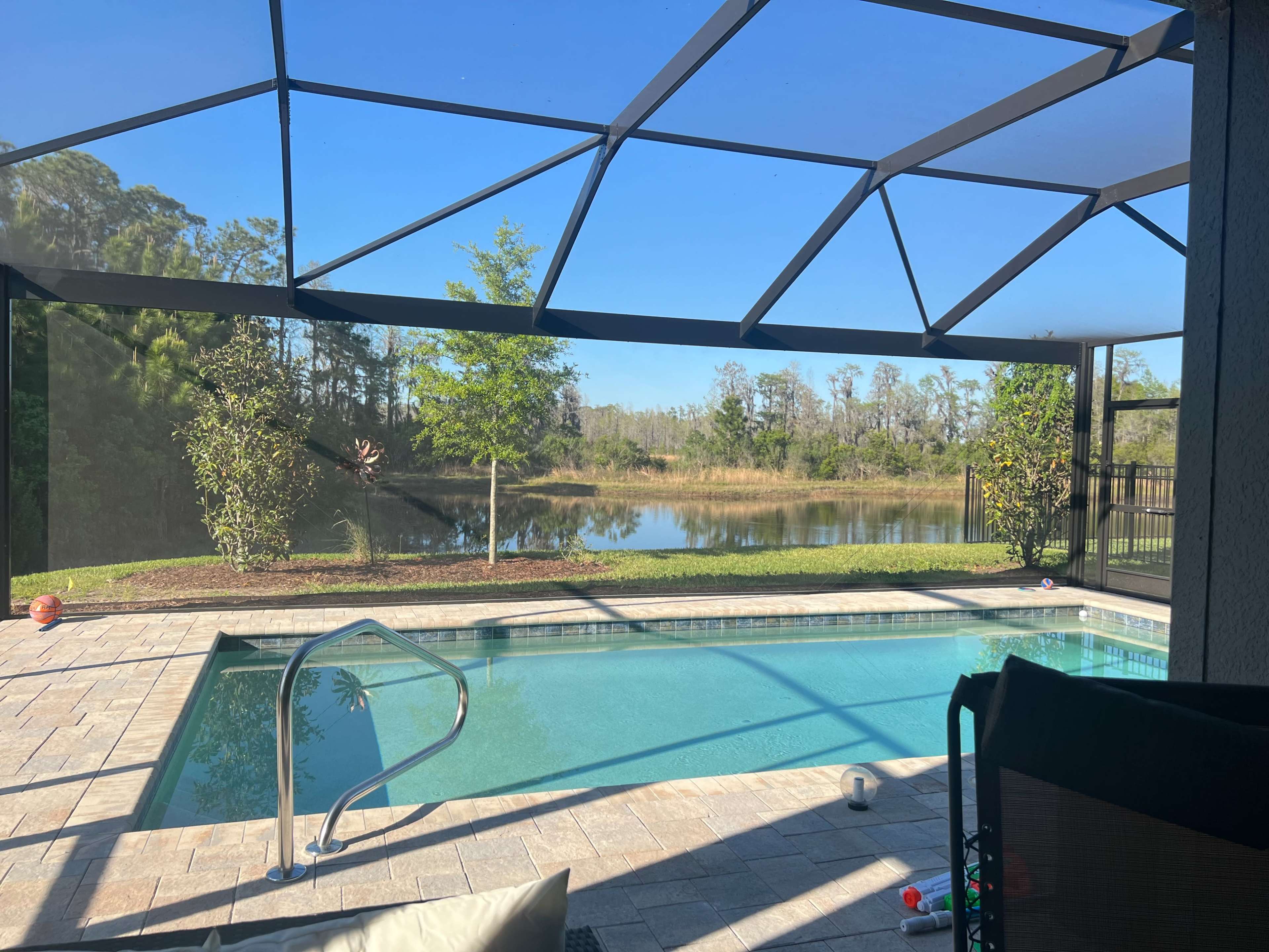 A swimming pool with a screened enclosure sits beside a lake, surrounded by green trees and a clear blue sky.
