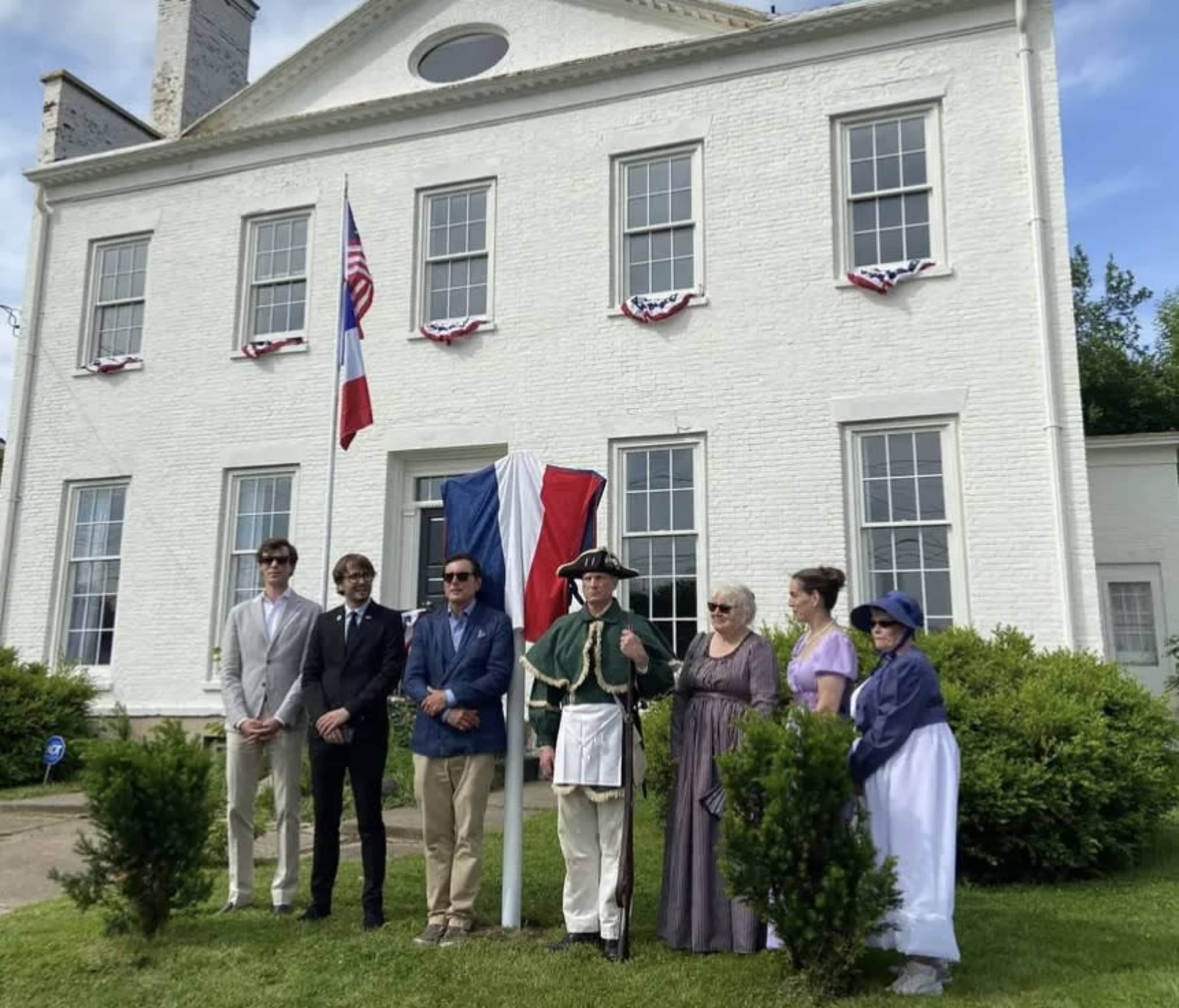 A group of people dressed in historical attire poses in front of a white building displaying flags and bunting.