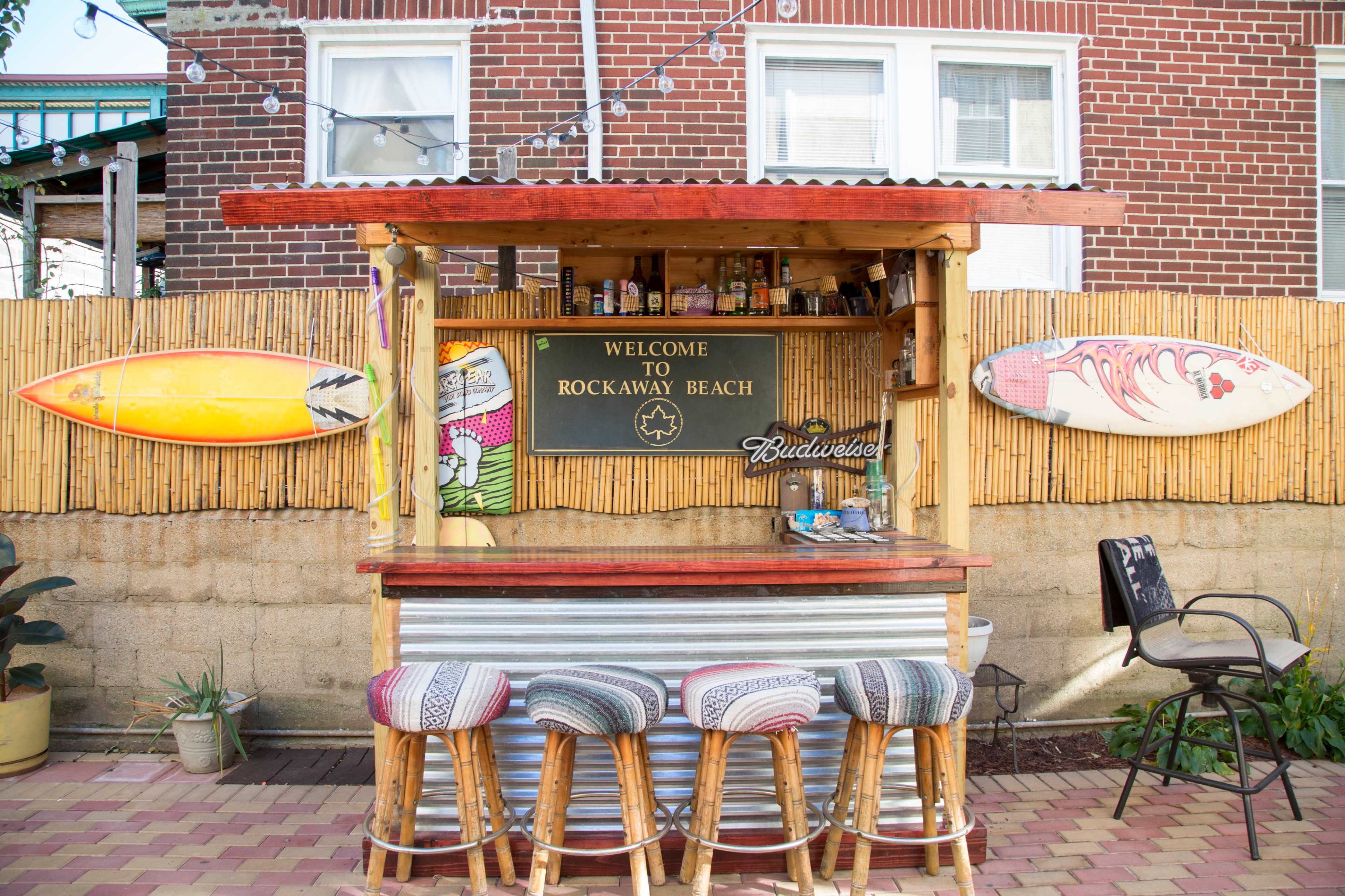A beach bar with a wooden counter and stools displays surfboards and a sign welcoming visitors to Rockaway Beach.