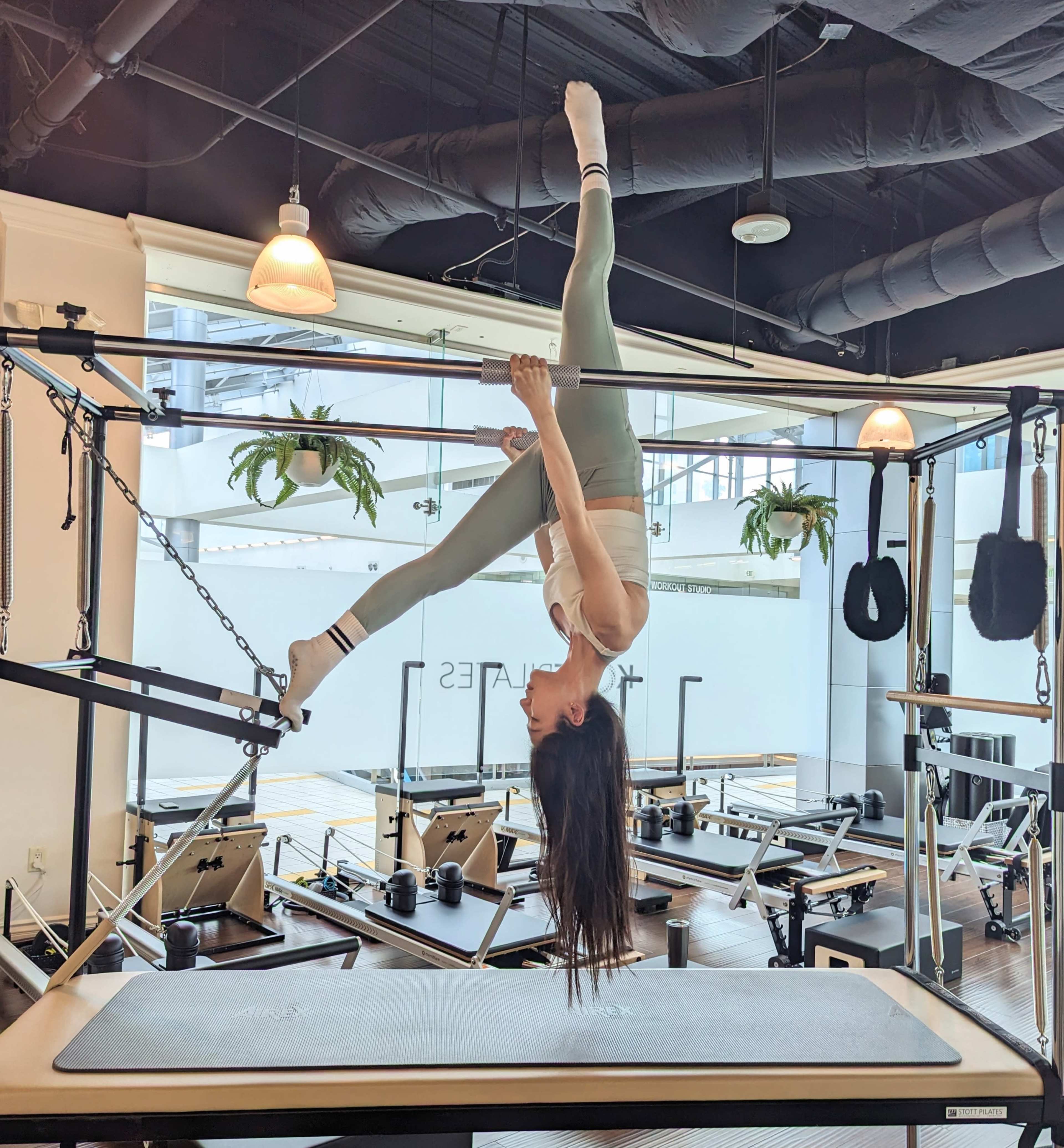 A woman is performing an inverted exercise on a Pilates reformer in a well-lit fitness studio.