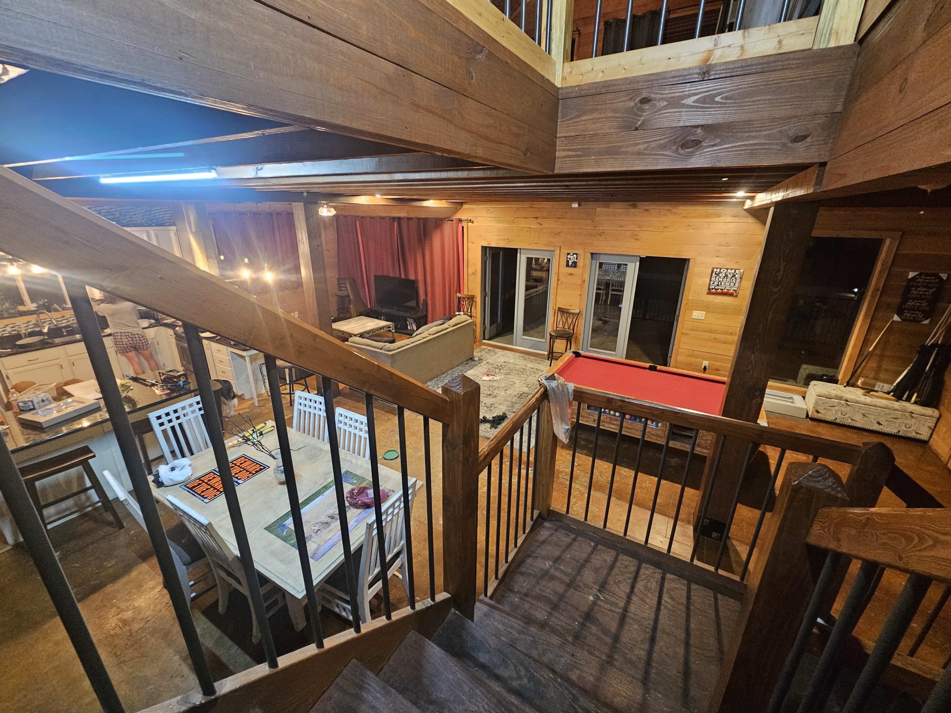 A multi-level wooden interior featuring a staircase leading to a balcony overlooking a living area with a dining table and a pool table.