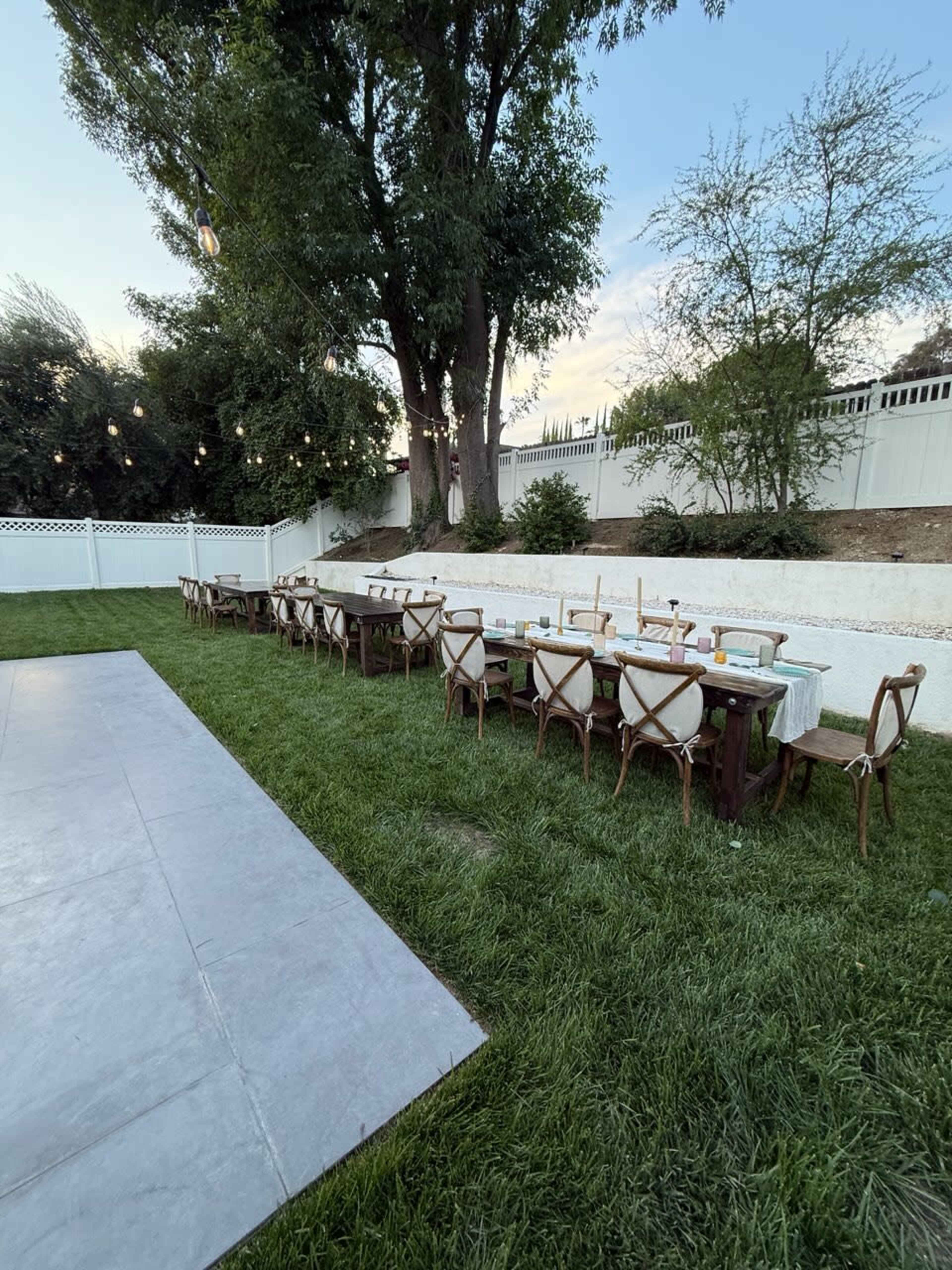A long wooden table with chairs is set up on a grassy area in a backyard, bordered by a white fence and illuminated by string lights above.