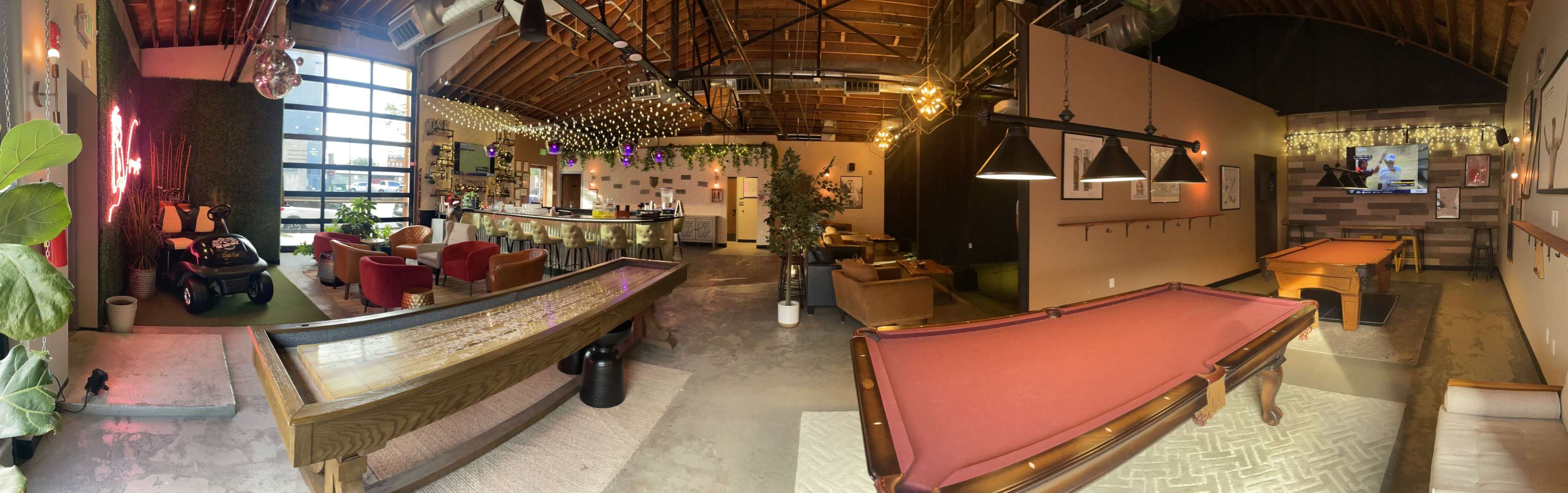 A spacious, modern lounge area featuring a bar, pool table, and various seating arrangements under a high ceiling with exposed beams.