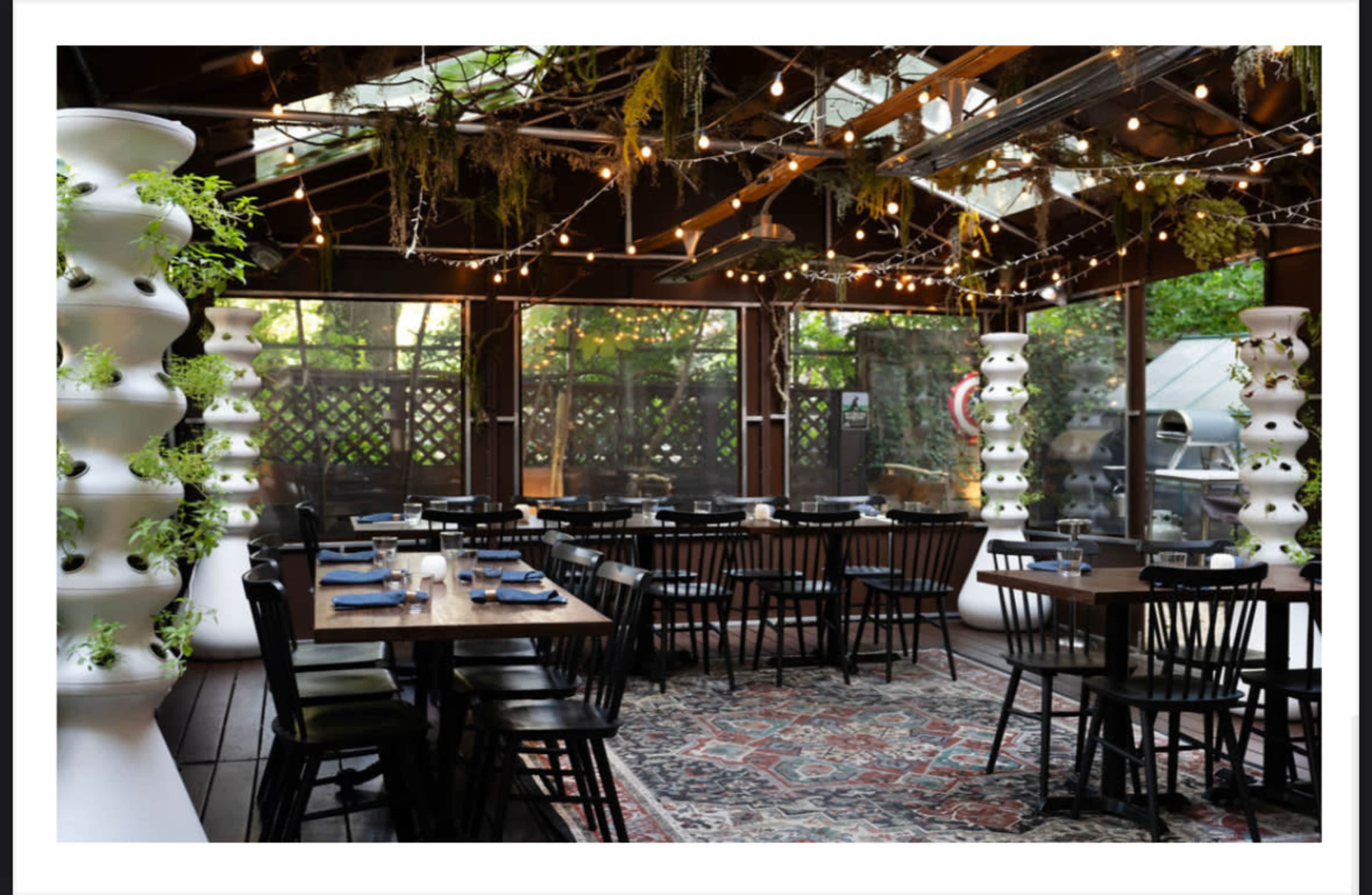 The image shows a restaurant interior with wooden tables, black chairs, and vertical planters adorned with greenery, illuminated by string lights.