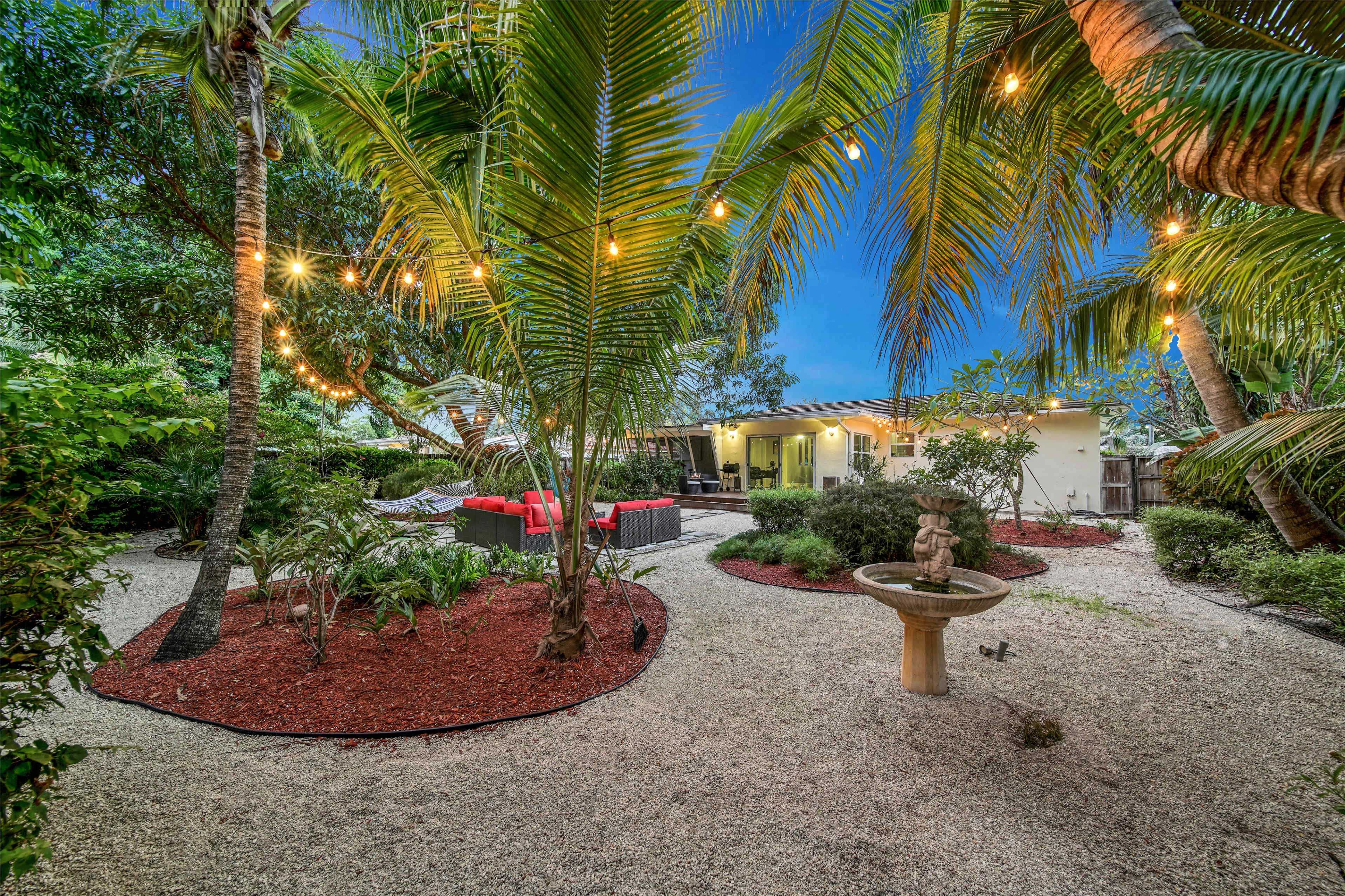 A lush backyard with palm trees, gravel pathways, a stone fountain, and a seating area illuminated by string lights.