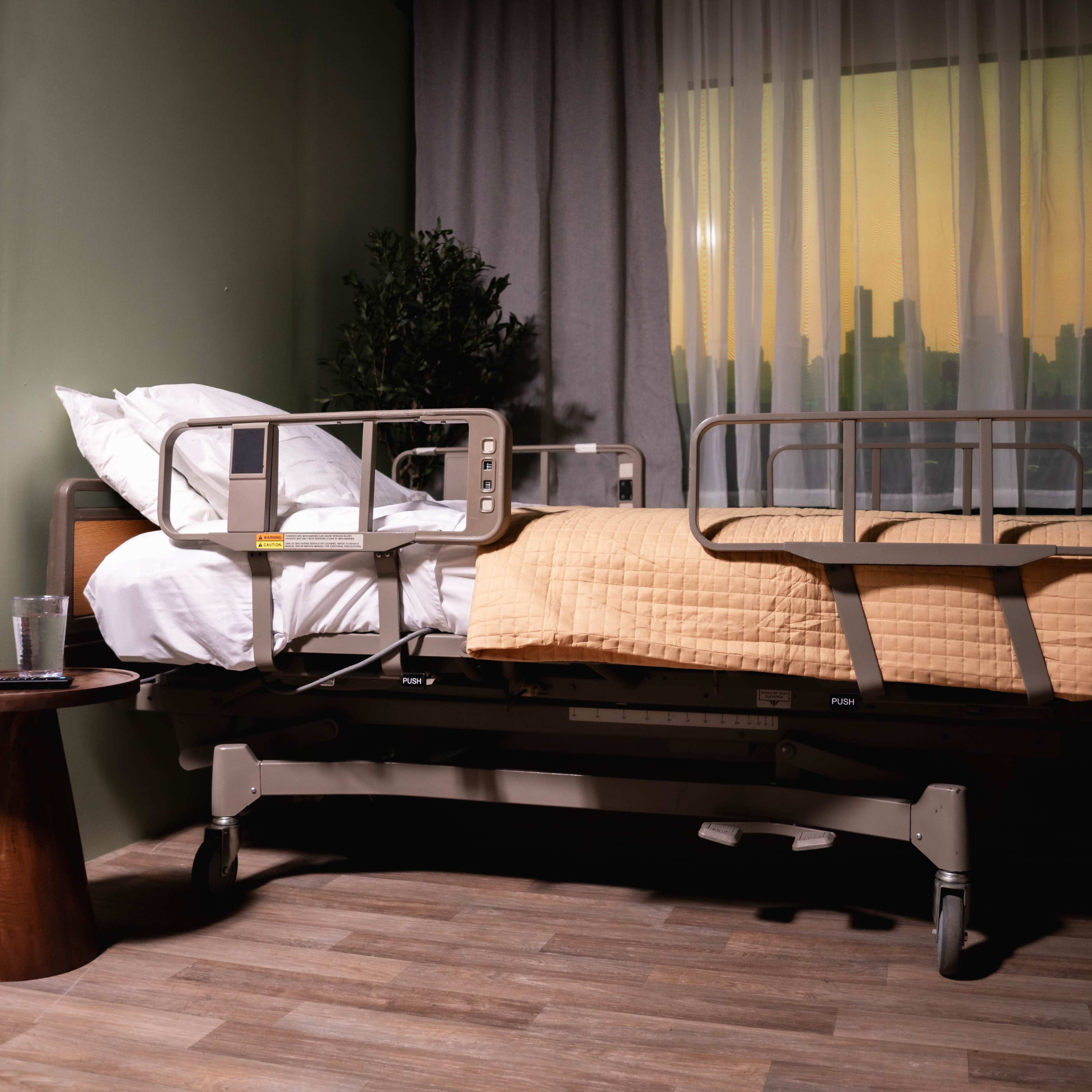 A hospital room features a bed with a yellow quilt and a bedside table next to it, set against a window showing a city skyline at sunset.