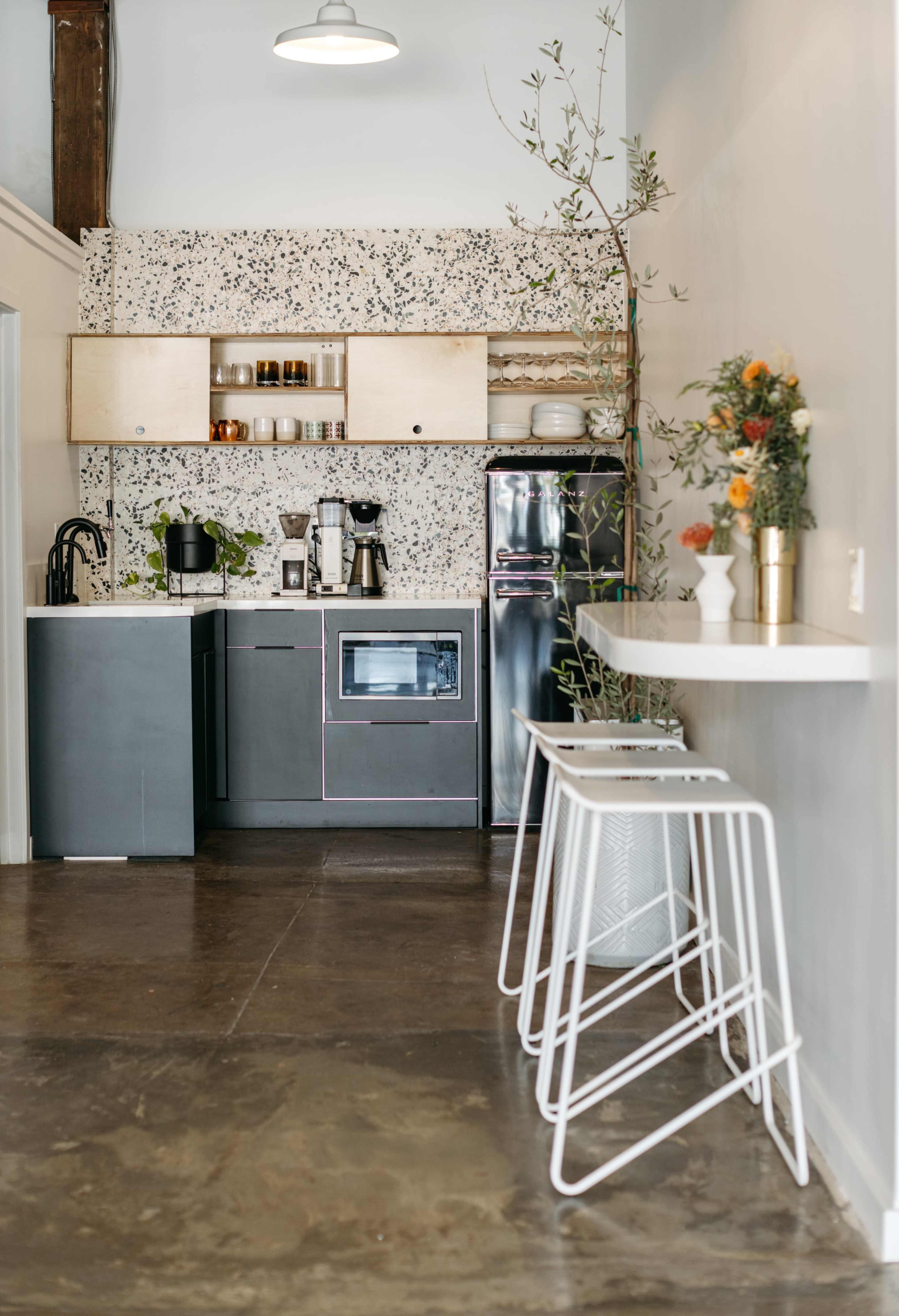 A modern kitchen features concrete floors, a speckled wall, and minimalist cabinetry with a coffee station set up.