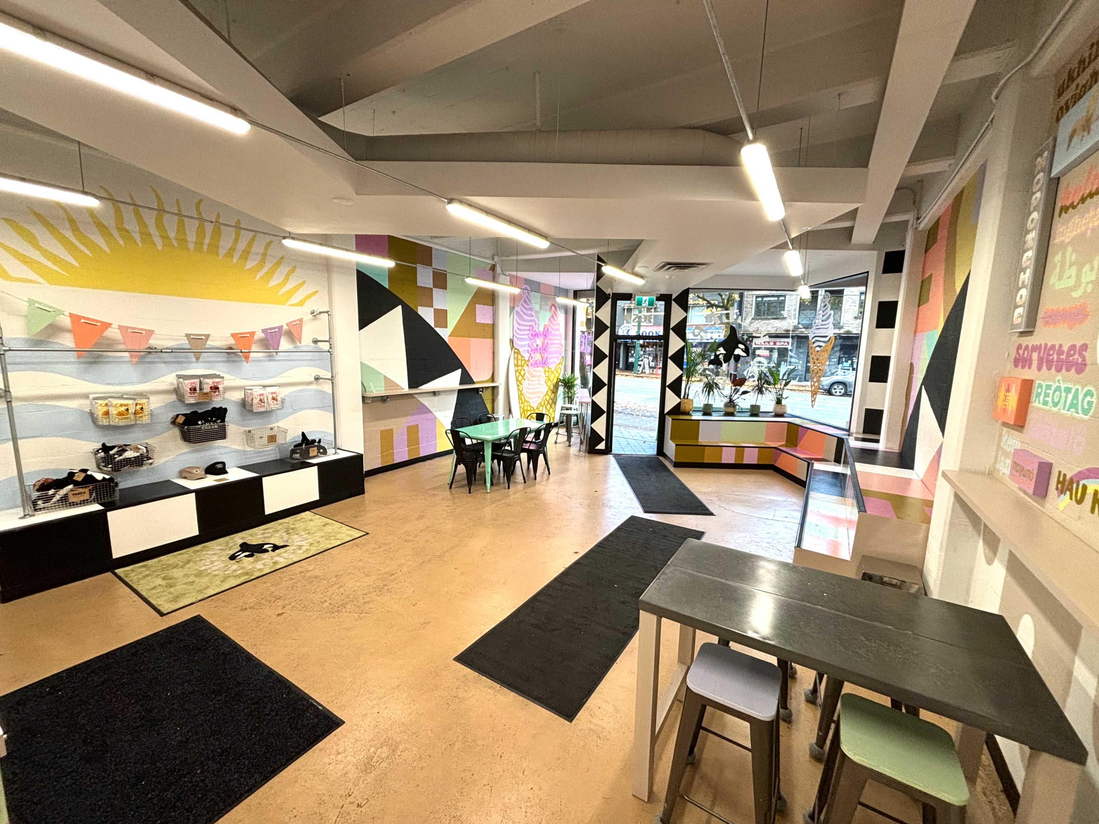 The image shows a colorful, modern interior of a storefront or café with geometric wall art, tables and chairs, and various decorative elements.