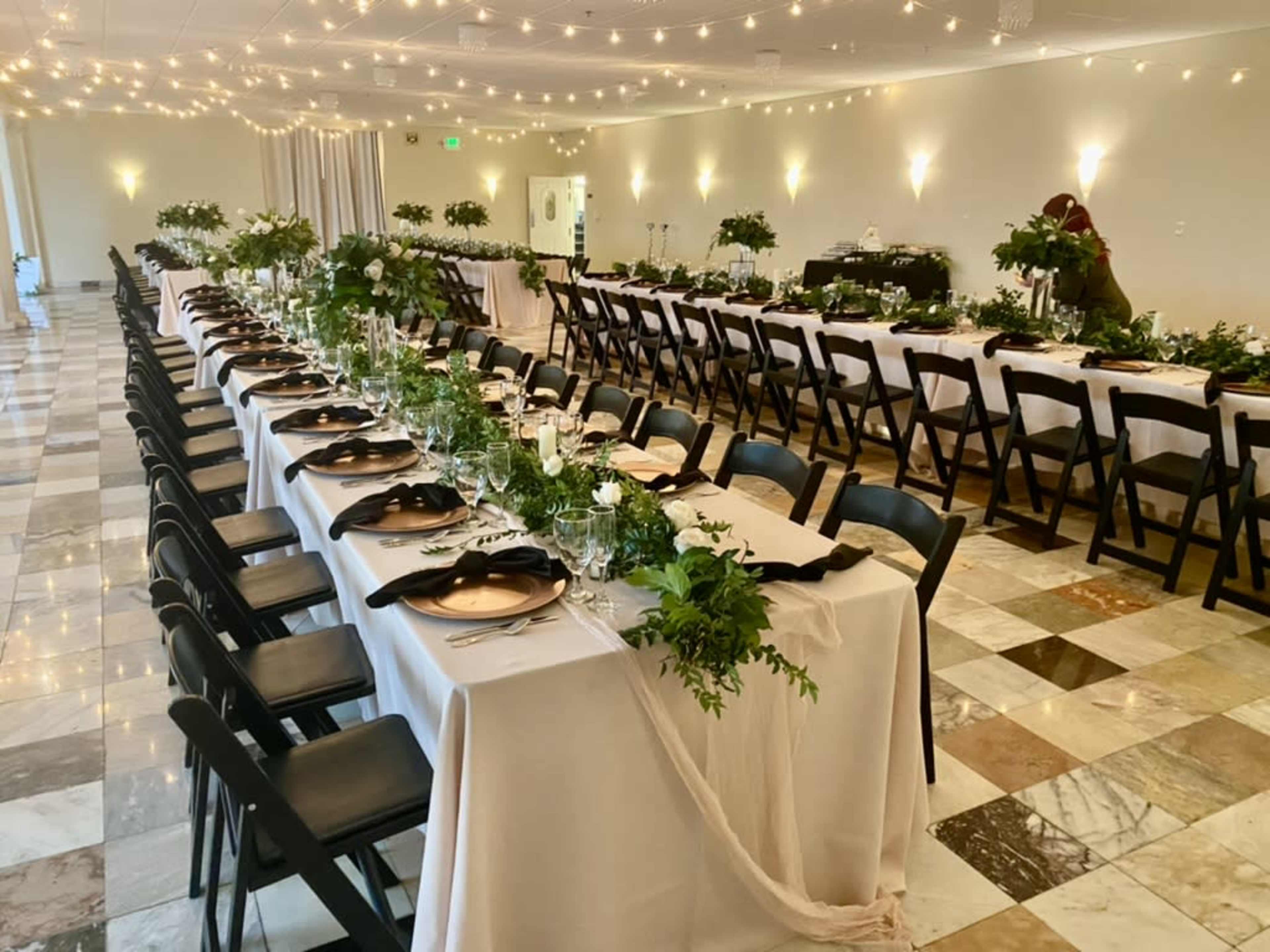 A spacious banquet hall features two long tables covered with white tablecloths, adorned with greenery and candles, set up for a formal event.