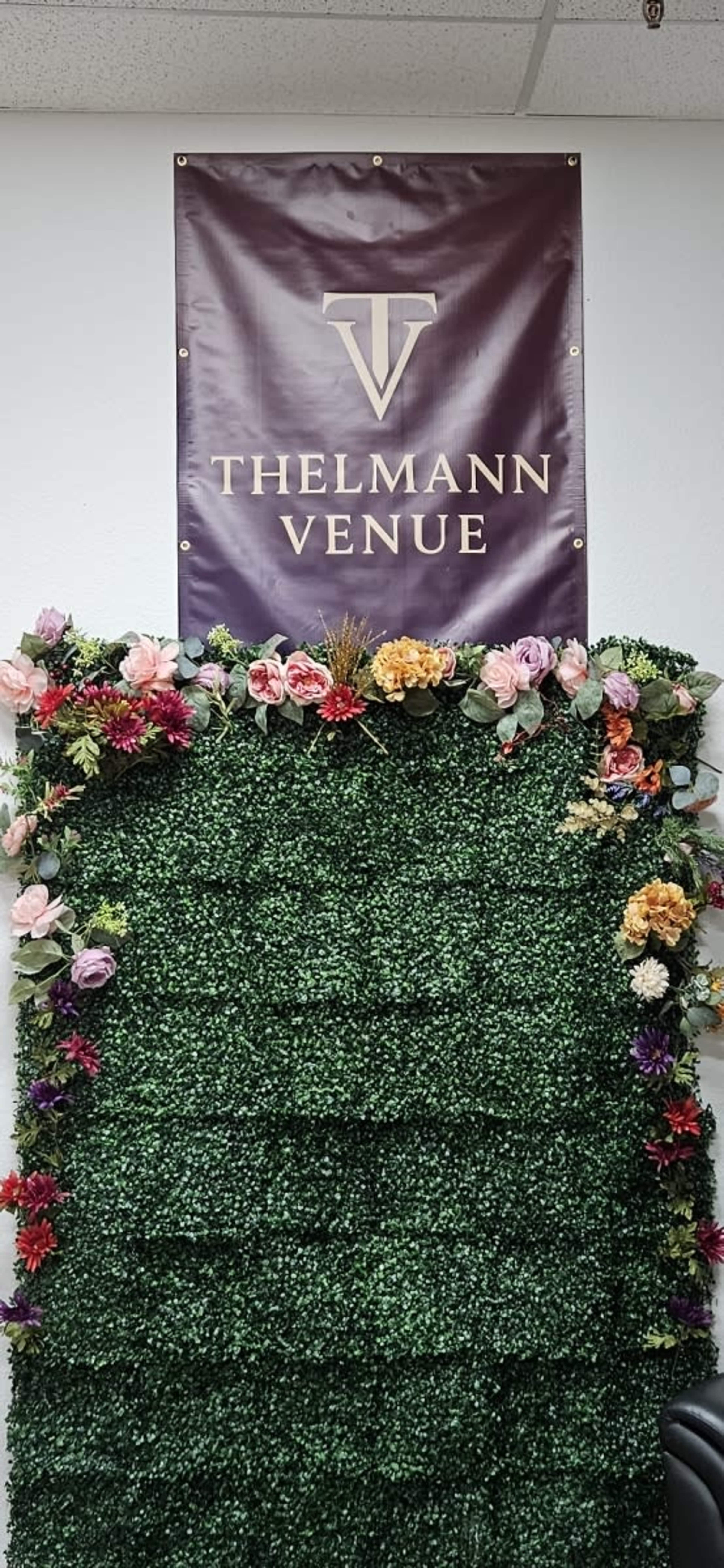 A wall adorned with artificial greenery and colorful flowers is topped by a sign that reads "THELMANN VENUE."