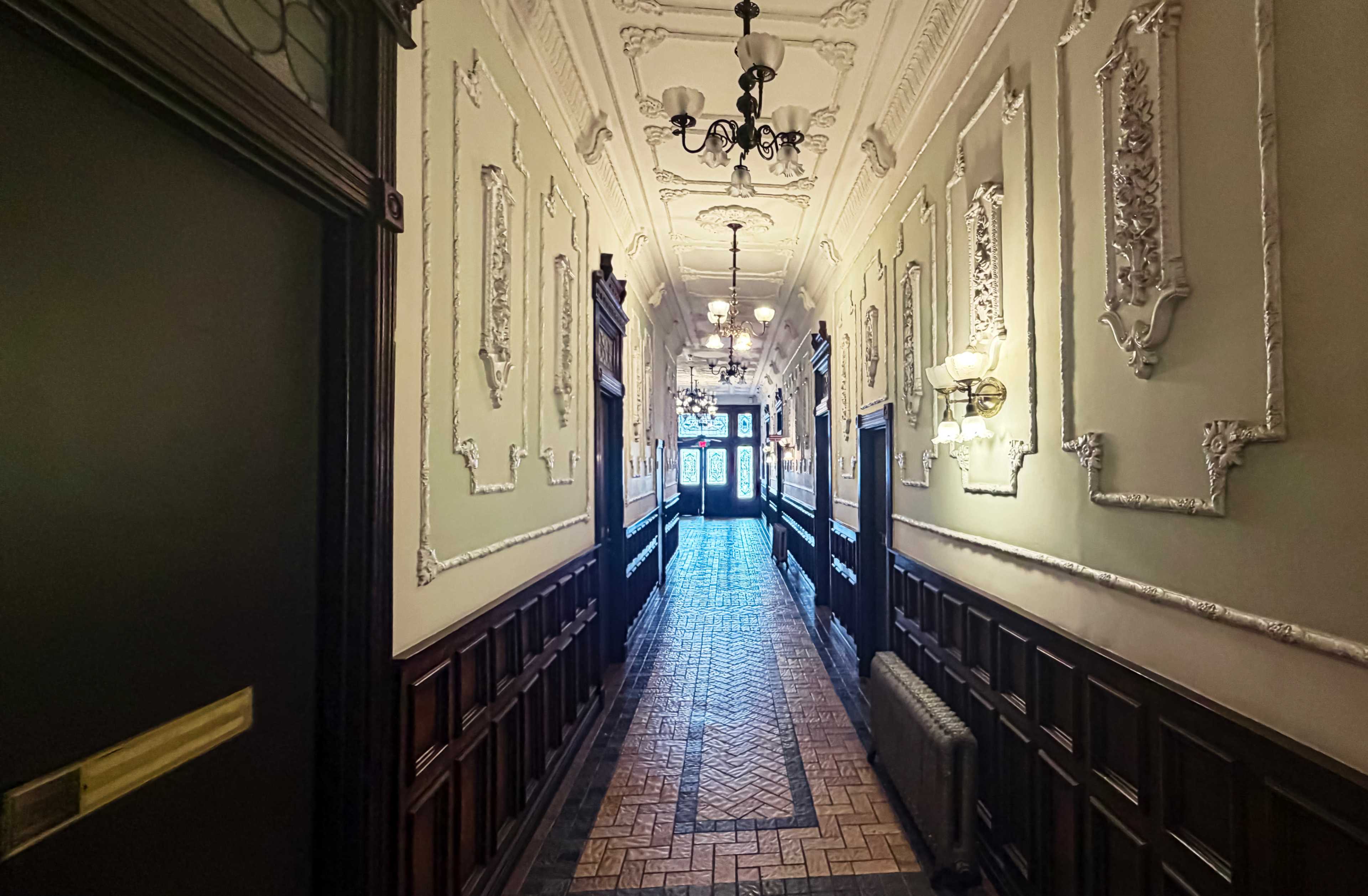 The image shows a long, elegantly decorated hallway with intricate moldings, chandeliers, and a patterned tile floor leading to a double door at the end.