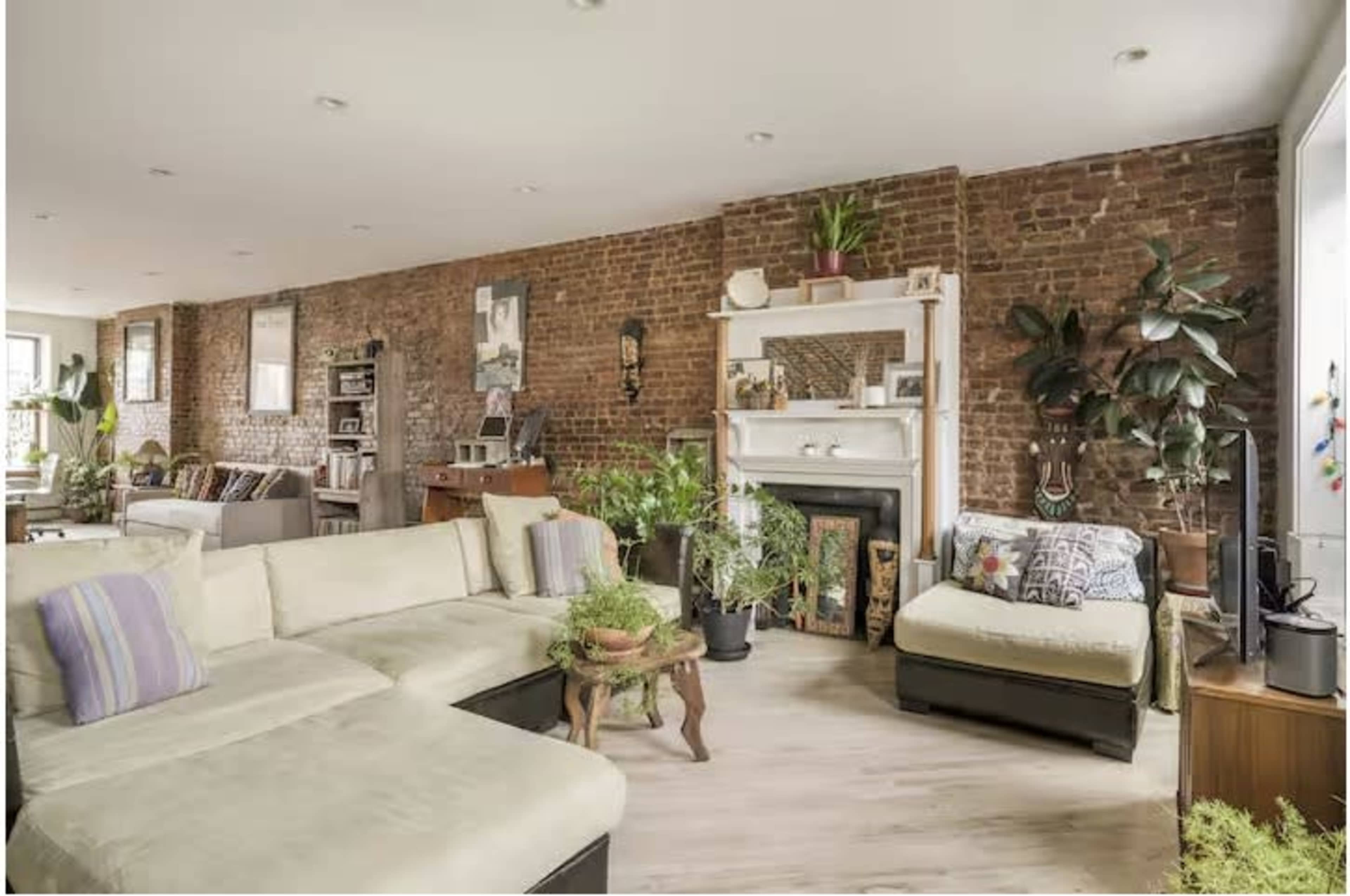 The image depicts a spacious, modern living room with exposed brick walls, featuring a sectional sofa, plants, and various decorative elements.