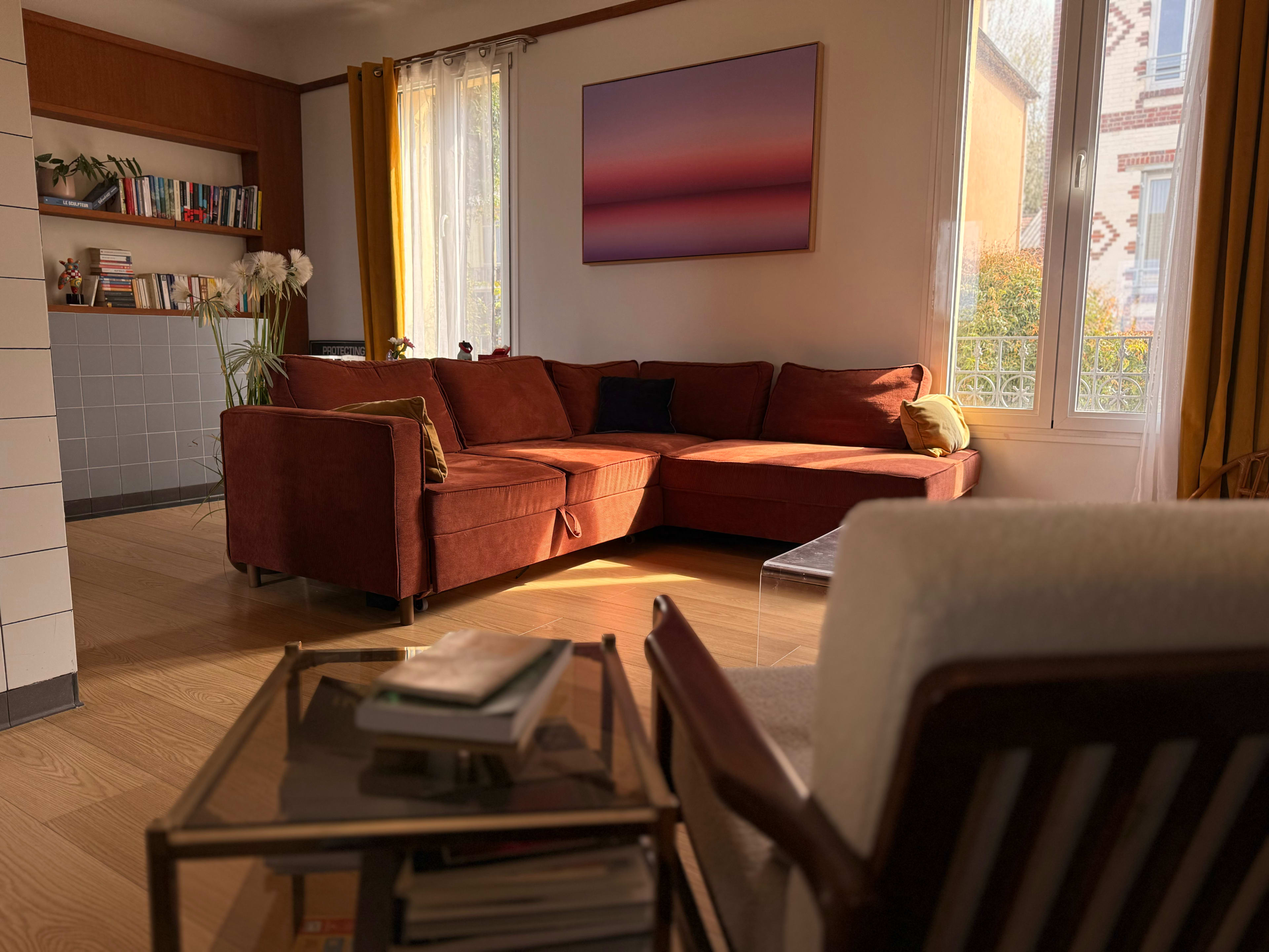 The image shows a cozy living room with a large brown sectional sofa, a glass coffee table, and a colorful wall art piece, illuminated by natural light through large windows.