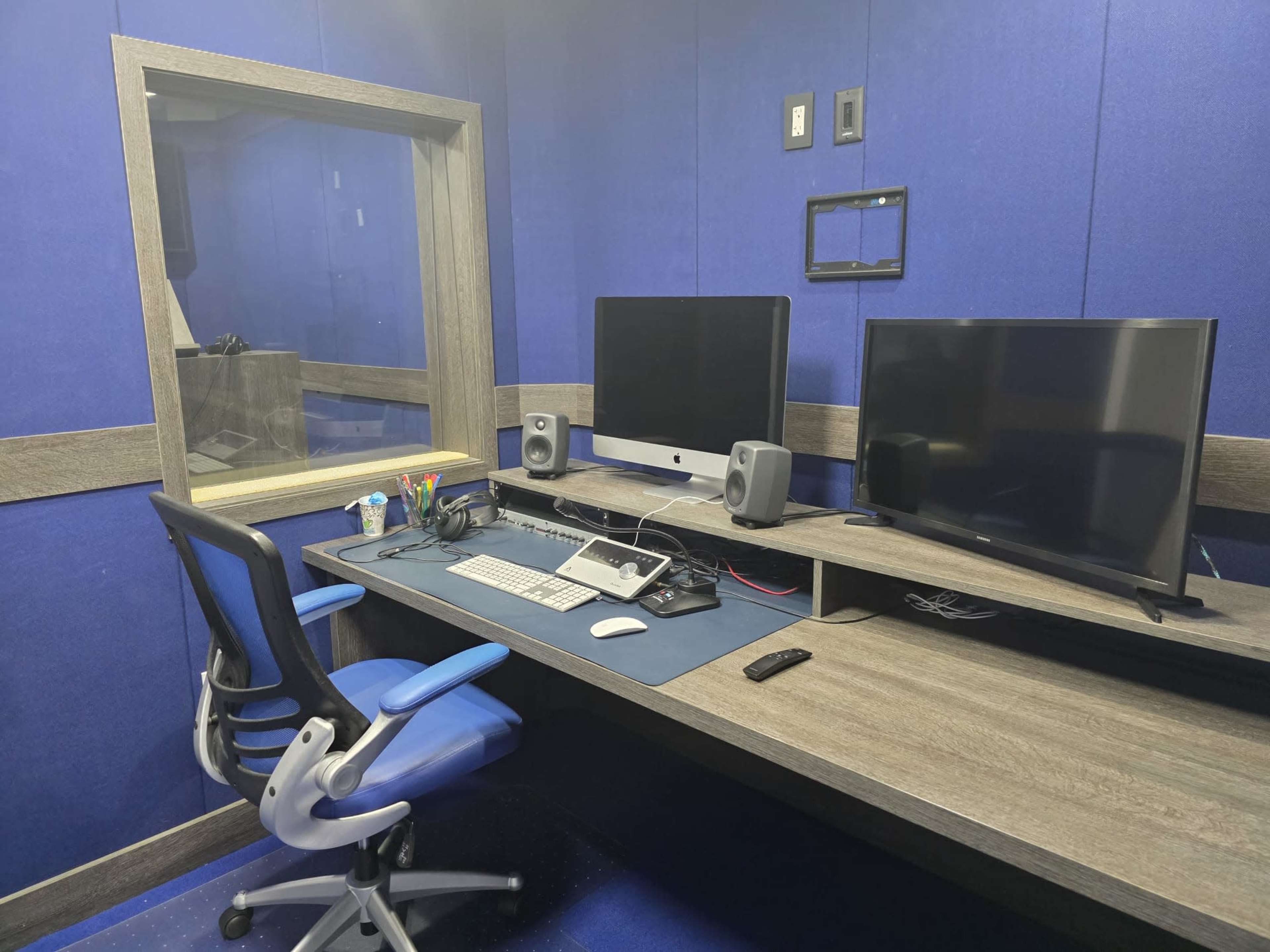 The image shows a small office space with a wooden desk, two computer monitors, a chair, and blue soundproof walls.