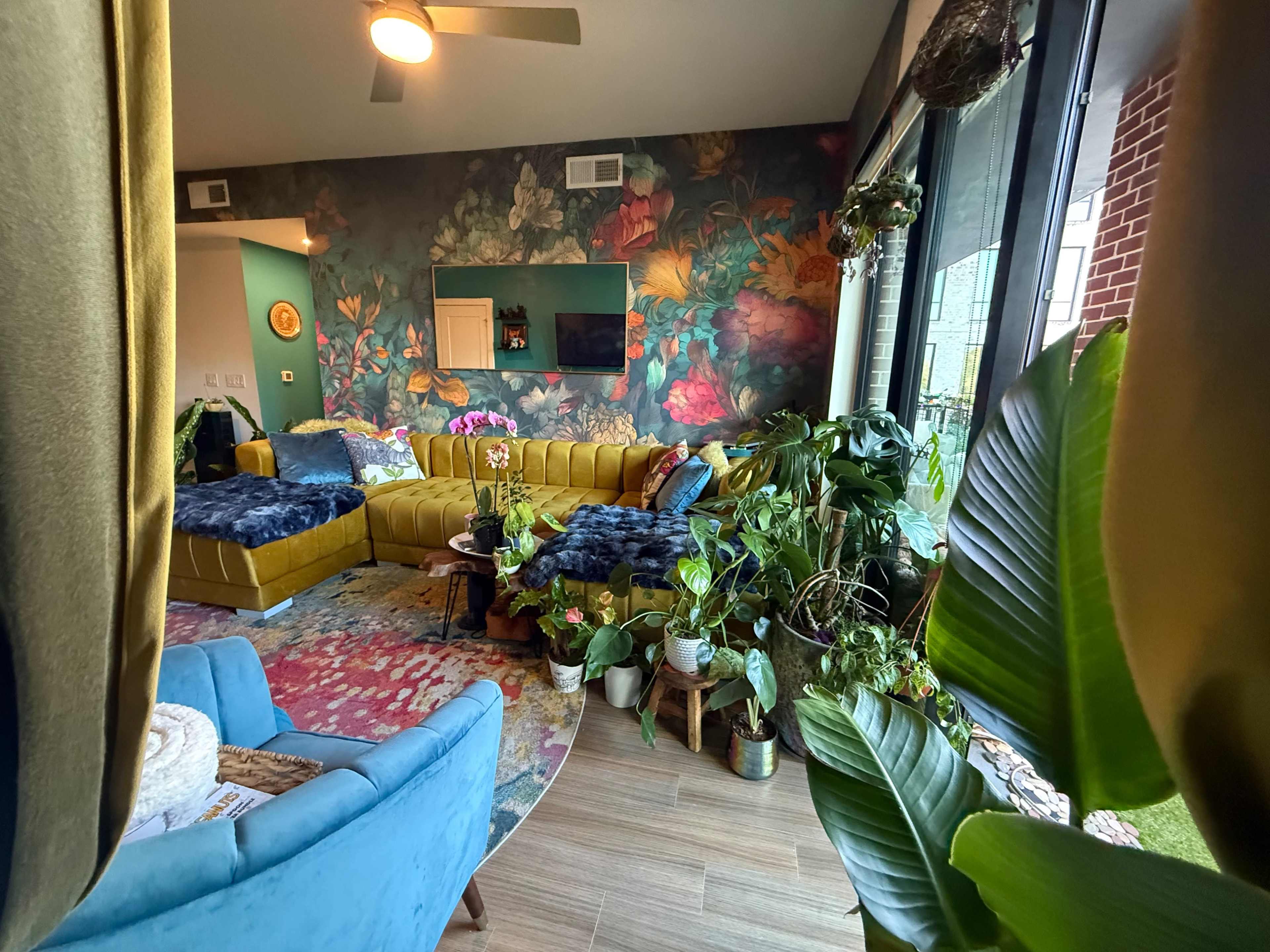 The image shows a vibrant living room featuring a large yellow sectional sofa, a decorative wall covered in floral wallpaper, and an assortment of indoor plants.