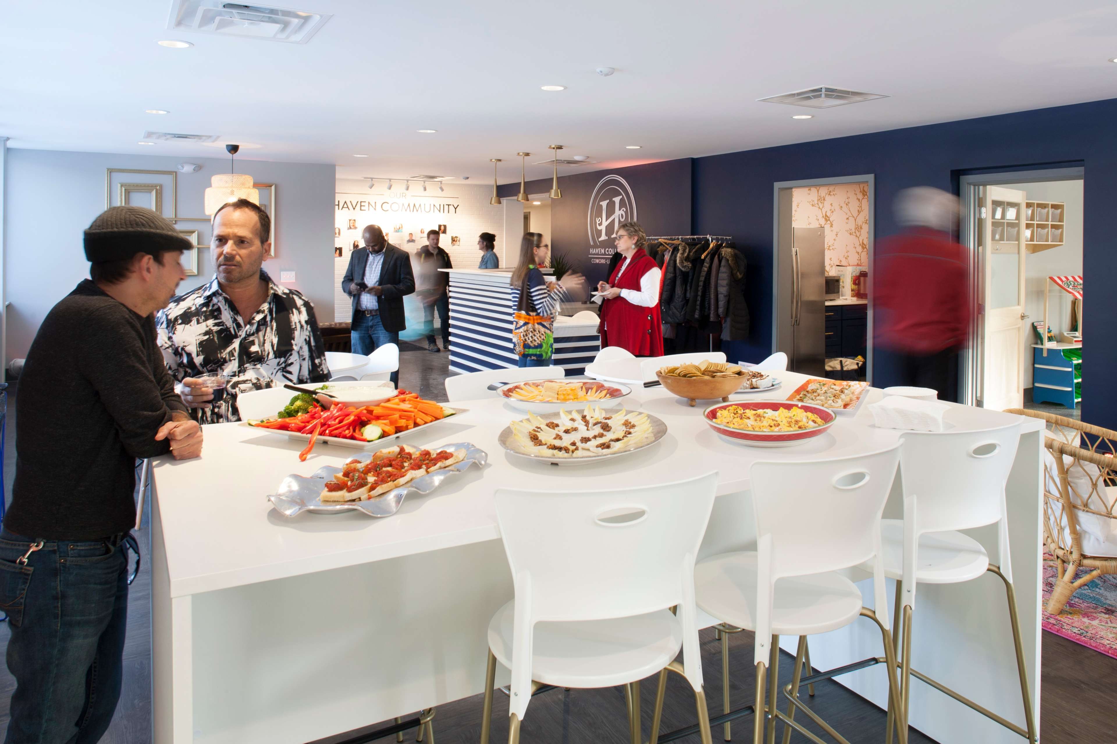 A social gathering in a modern space with people engaging in conversation around a white table filled with various food platters.