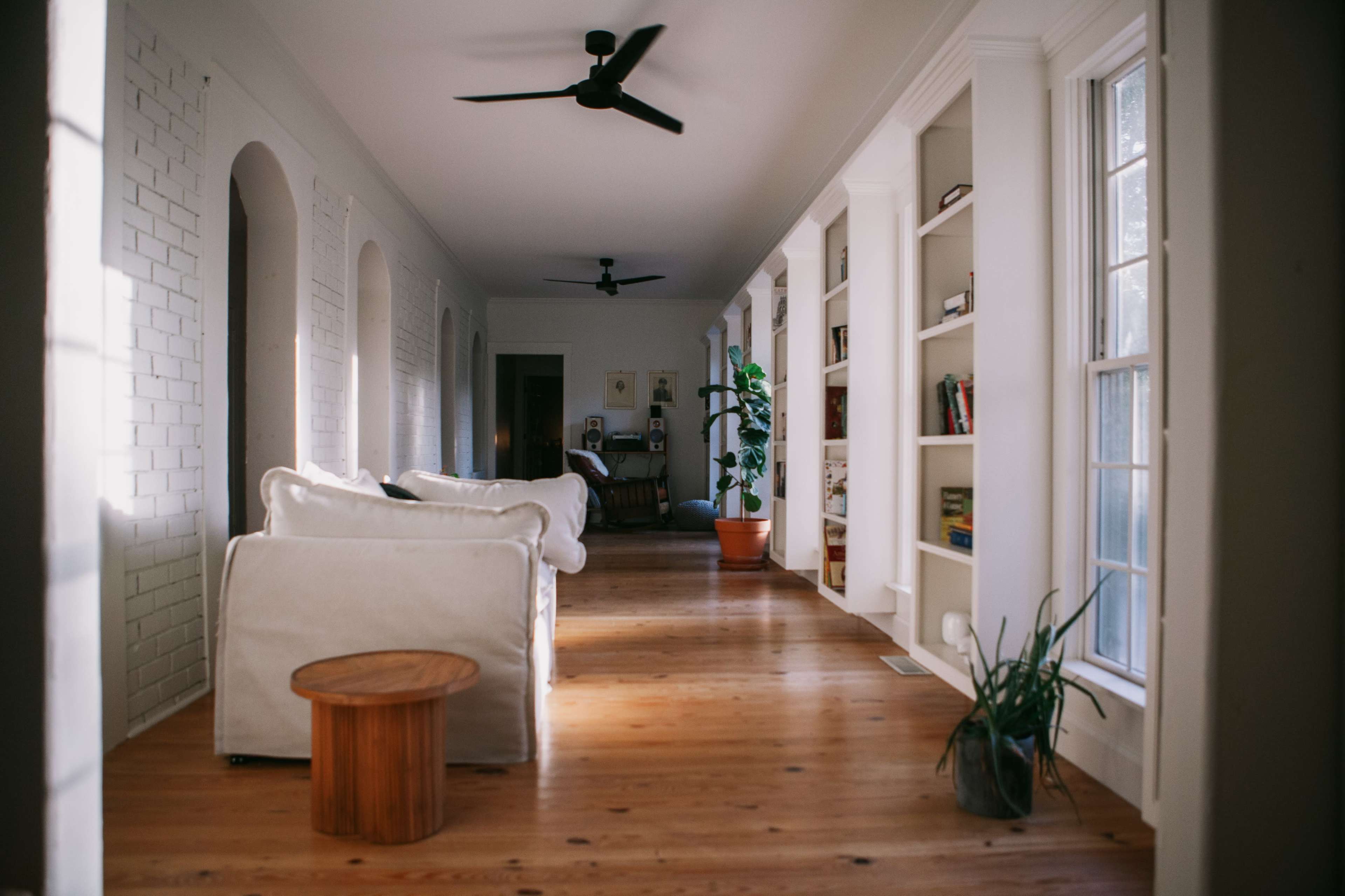 A bright corridor features a white sofa, wooden stool, bookshelves, and large windows, illuminated by natural light.