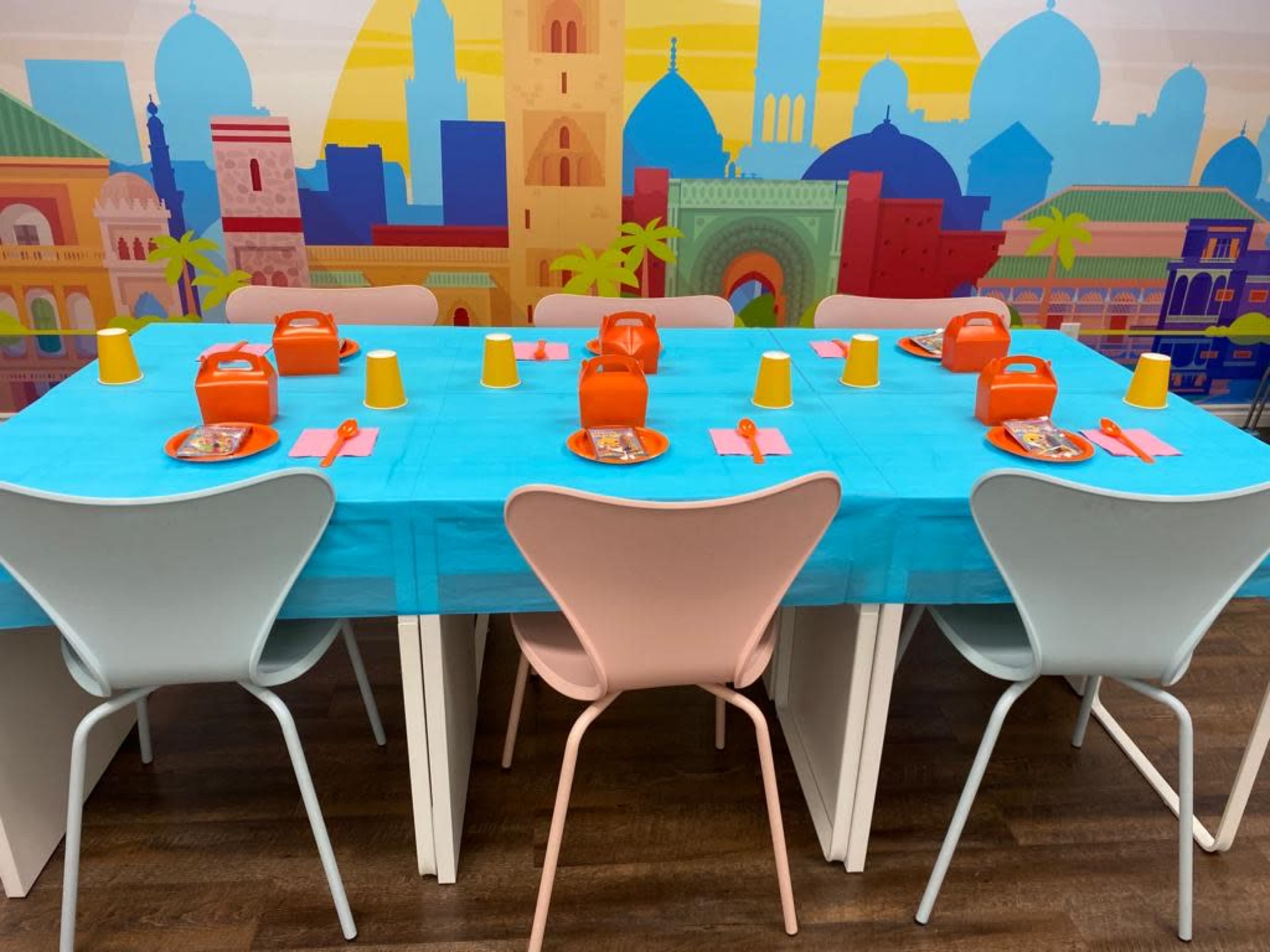 A dining table is set with blue tablecloths, orange plates, yellow cups, and pink utensils, against a colorful background featuring cityscapes and palm trees.