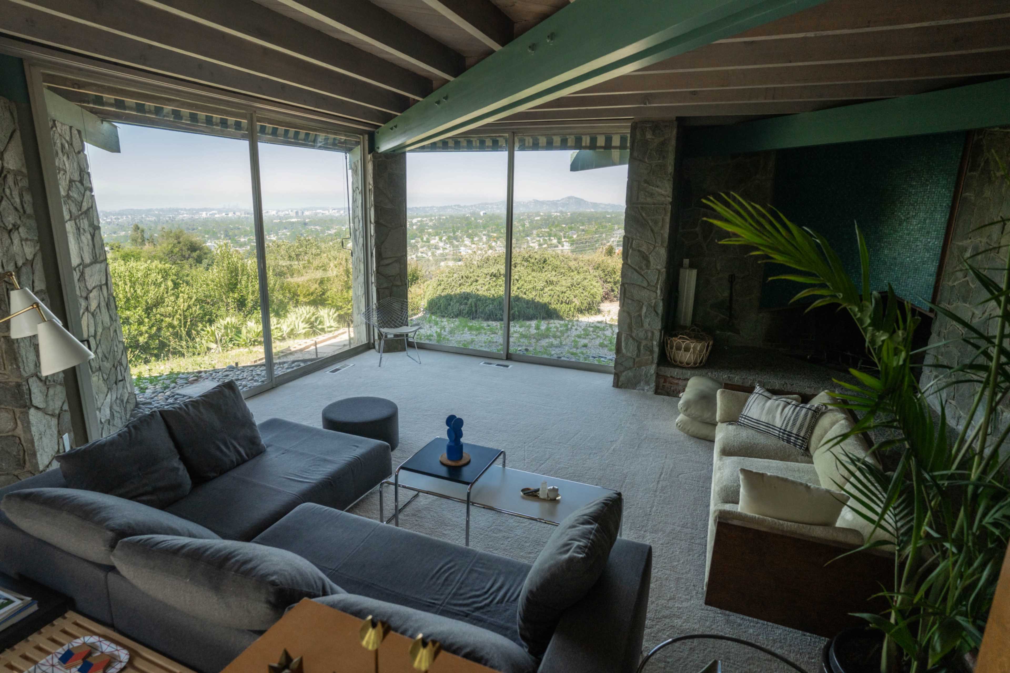 The image shows a modern living room with large windows offering a panoramic view of the landscape, furnished with a gray sofa, a white chair, and a coffee table.