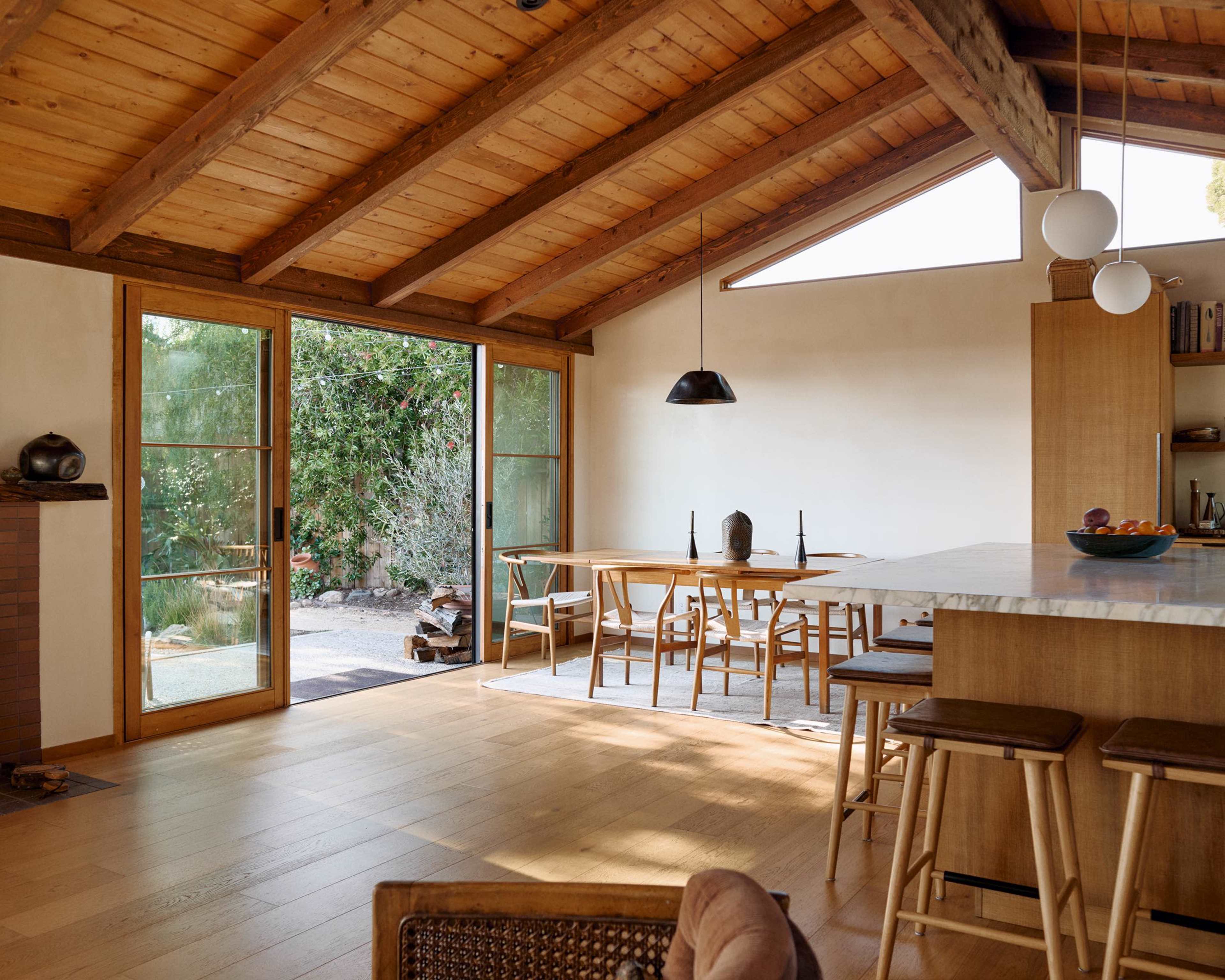 The image shows a modern interior of a wooden-beamed living space with a dining area and a kitchen island, illuminated by natural light from large windows.