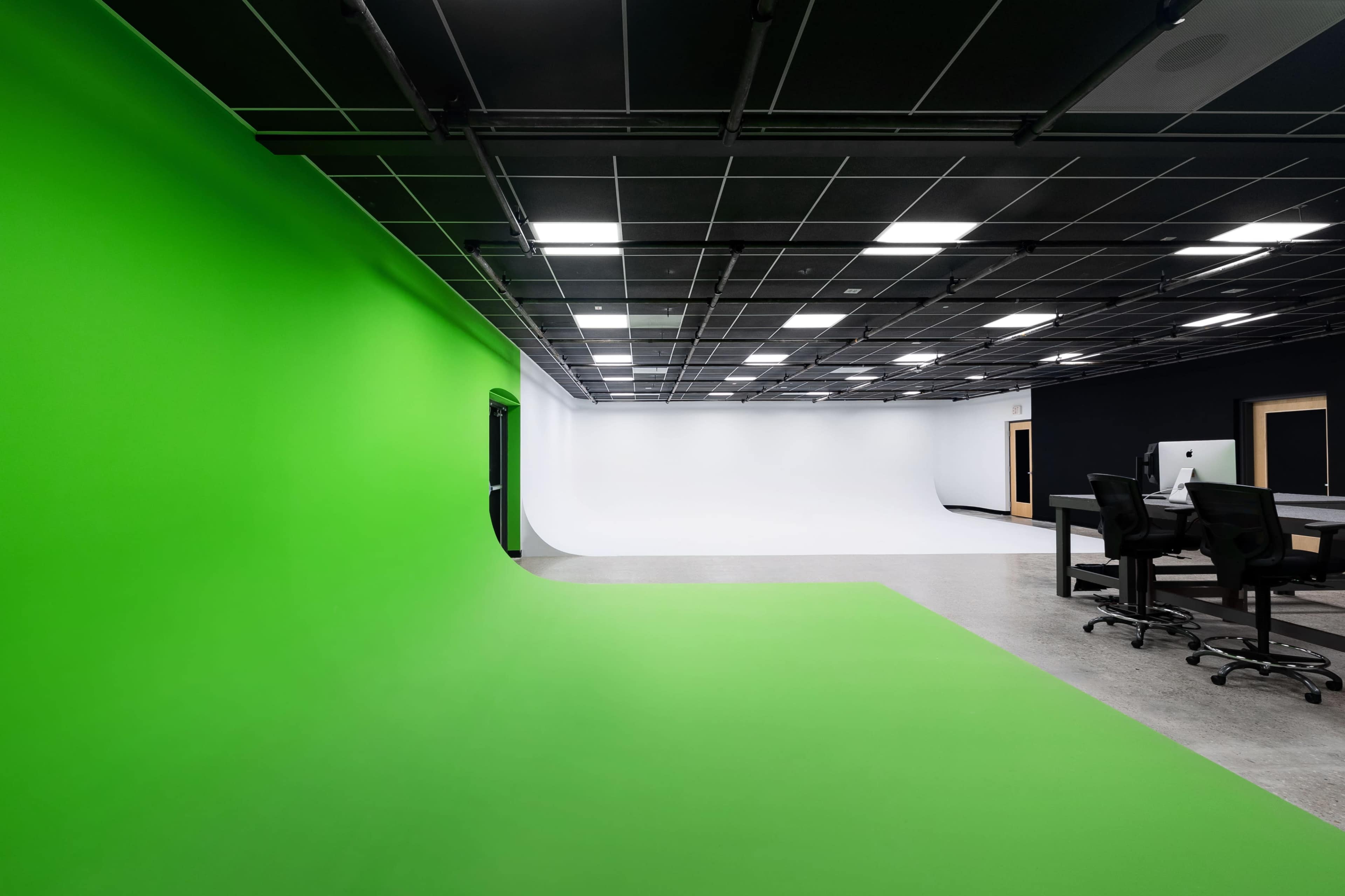 A spacious studio with a green backdrop on one side and a white backdrop on the opposite side, featuring a black wall and a work desk with chairs.
