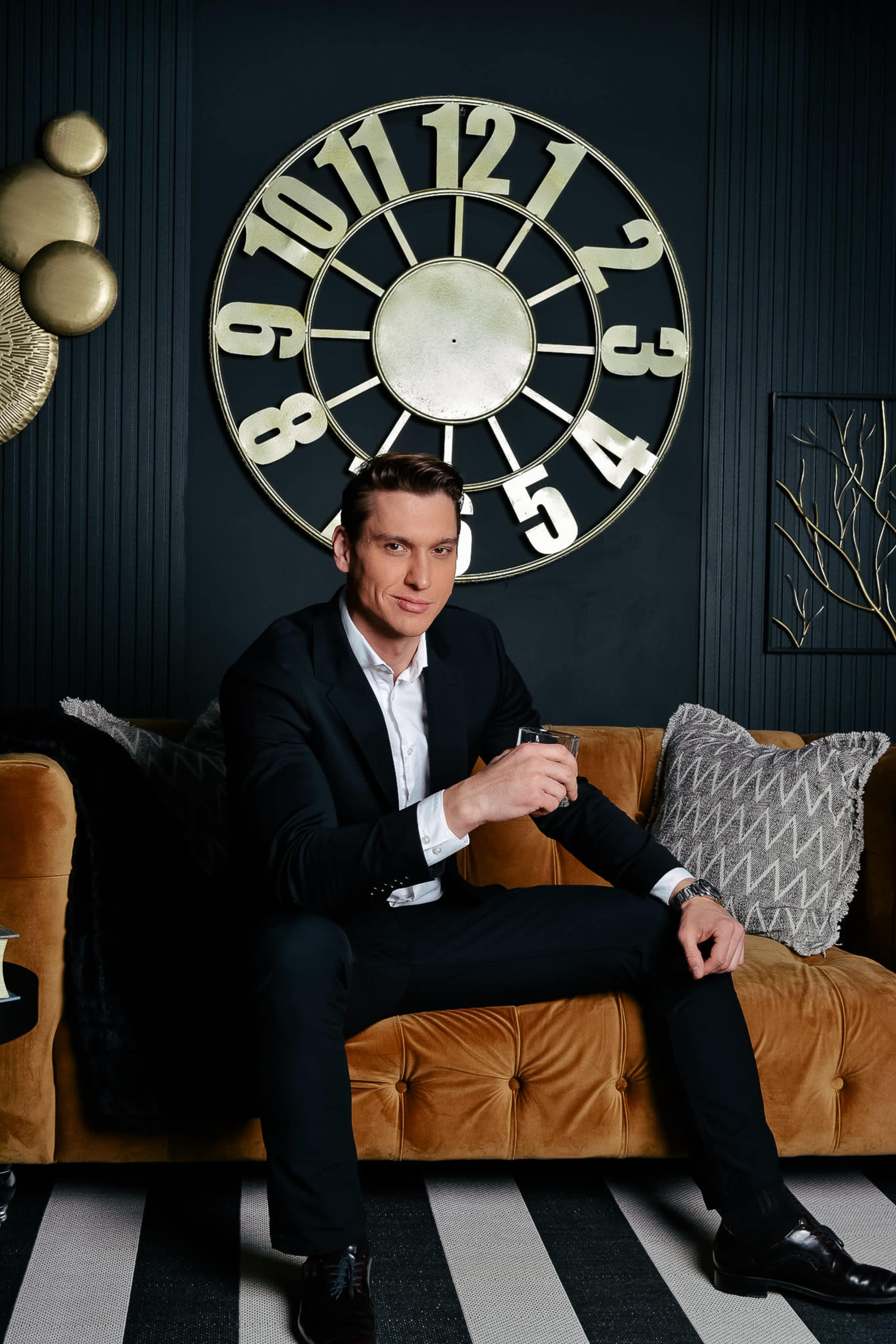 A man in a black suit sits casually on a tan sofa, holding a drink, against a backdrop featuring a large wall clock.