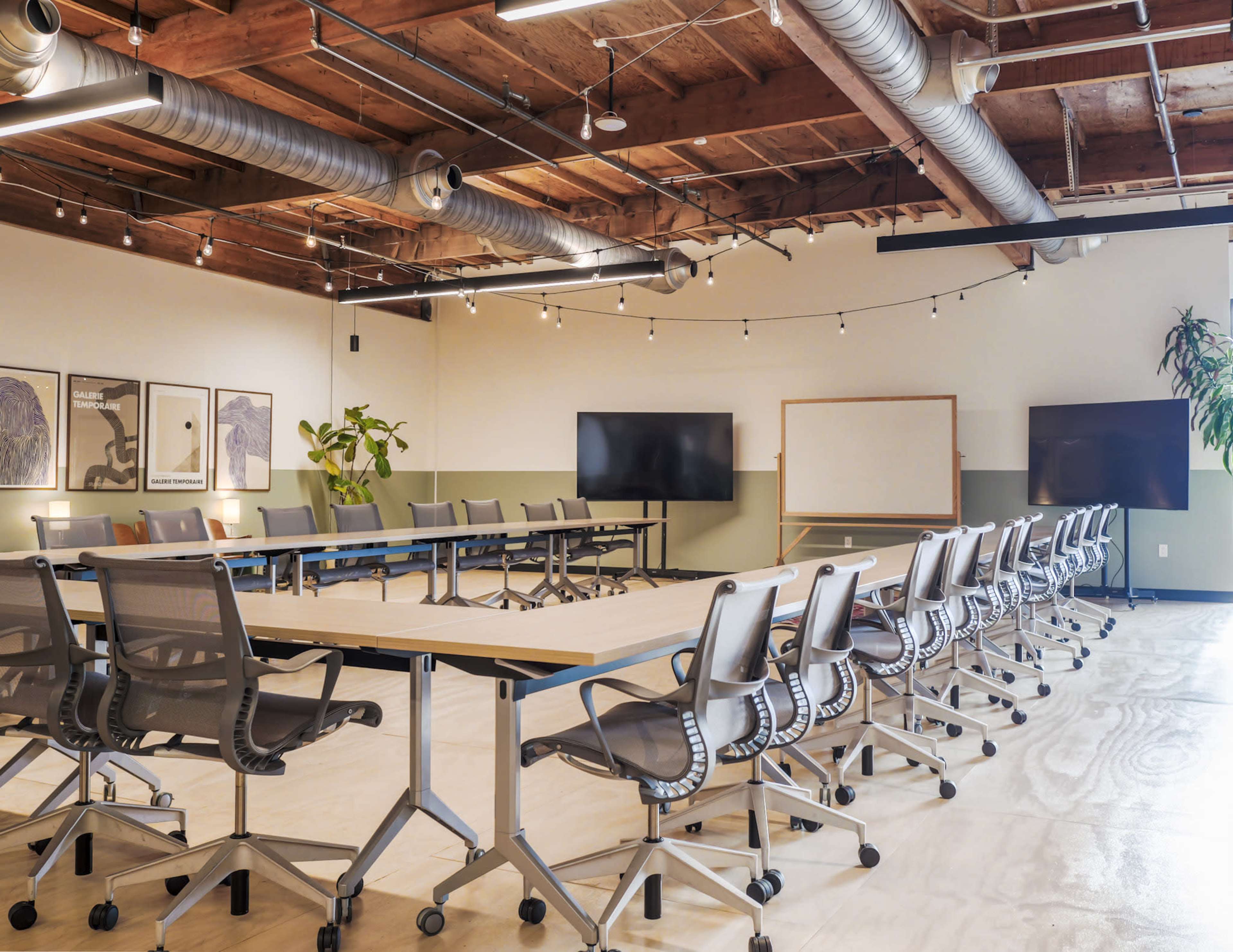 The image shows a modern conference room equipped with multiple tables arranged in a U-shape, ergonomic chairs, and large screens on the walls.
