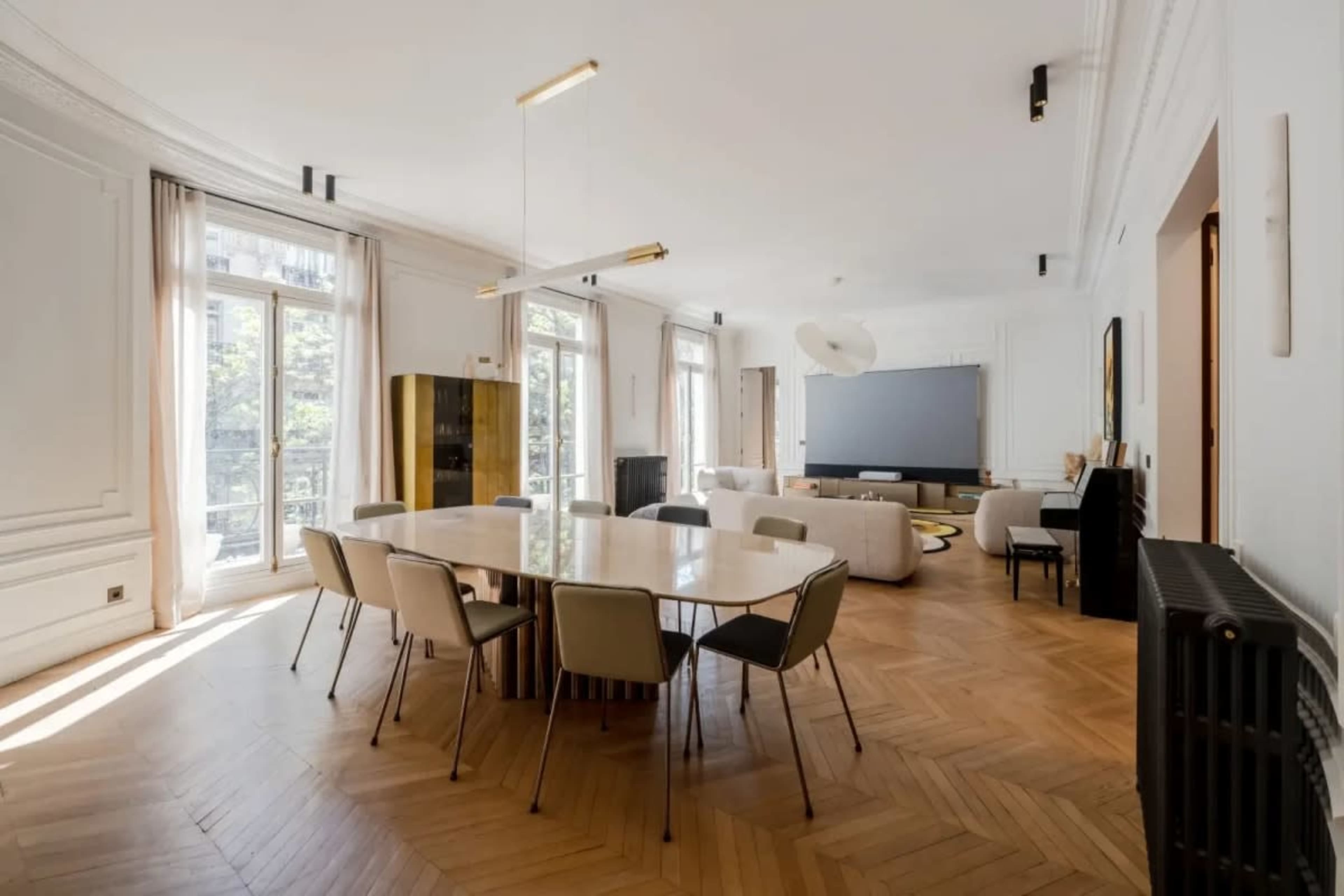 The image shows a spacious, well-lit living area with a large dining table, modern furniture, and floor-to-ceiling windows.