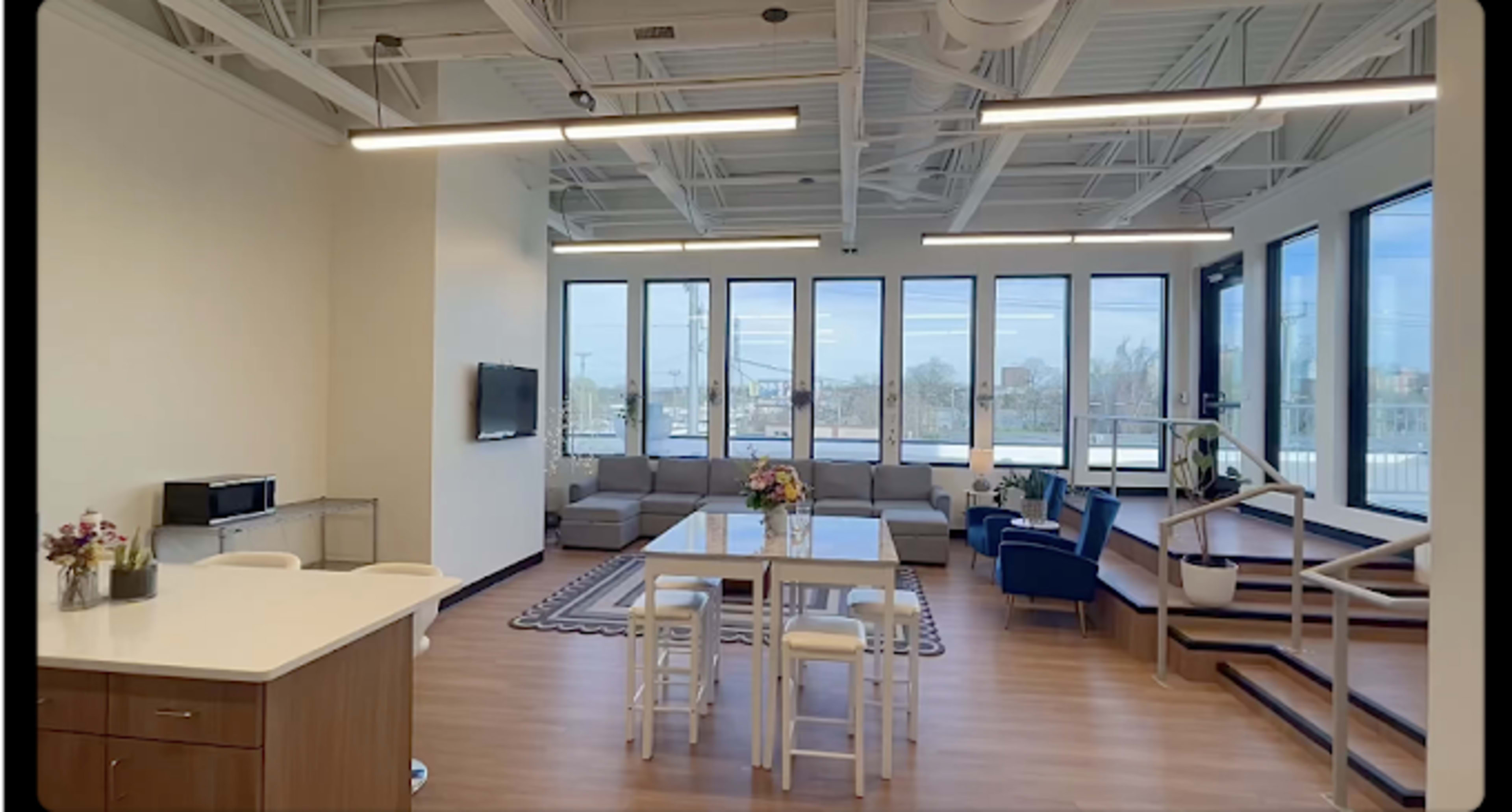The image shows a modern, bright office space with large windows, a dining table, chairs, and a staircase leading to a different level.