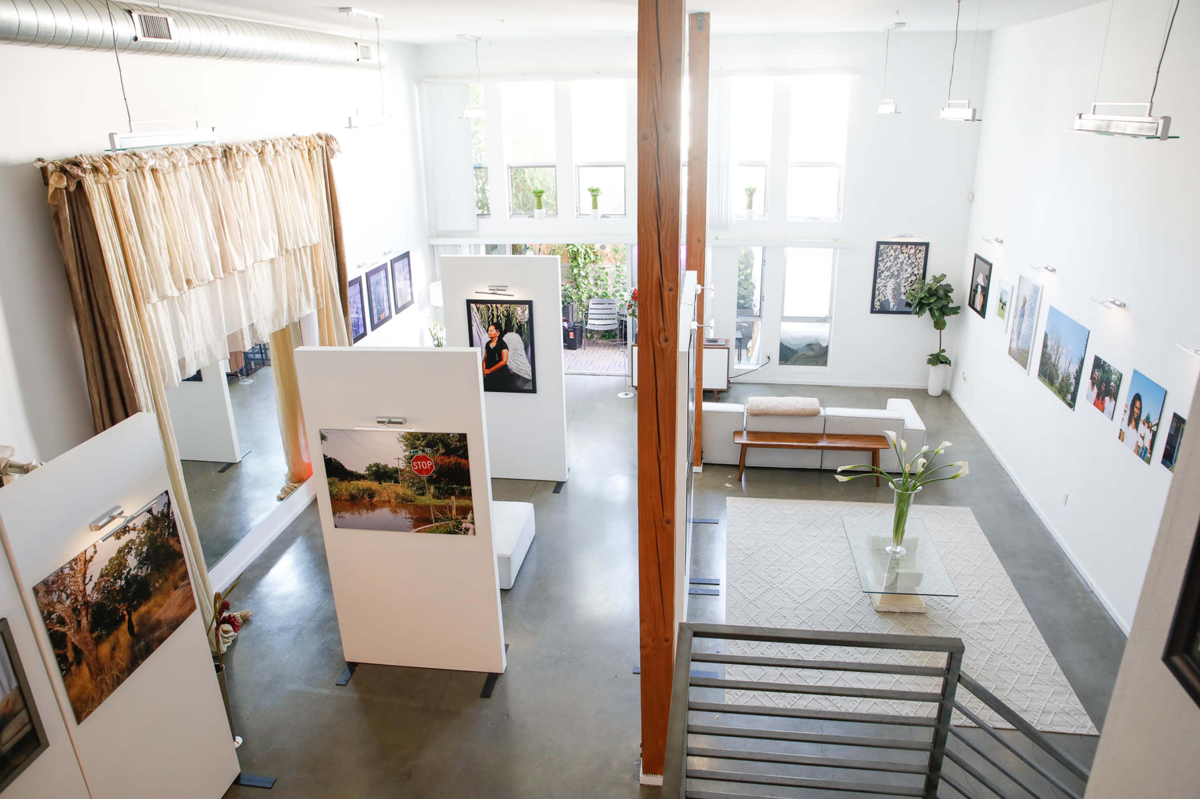 A spacious art gallery with white walls, tall windows, and exhibition panels displaying photographs surrounded by a modern seating area.