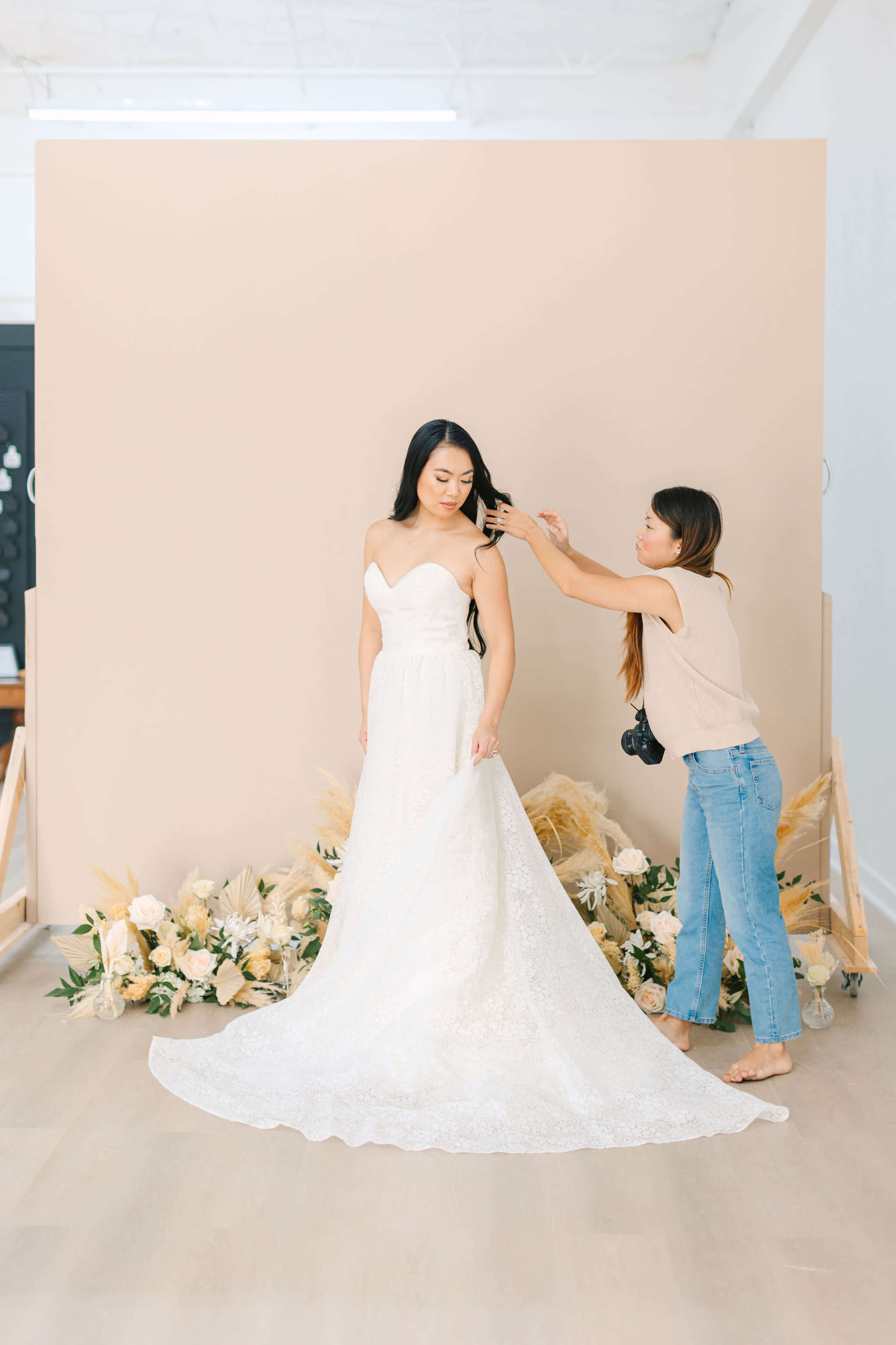 A woman in a wedding dress stands next to a stylist who is adjusting her hair in front of a neutral backdrop adorned with flowers and greenery.