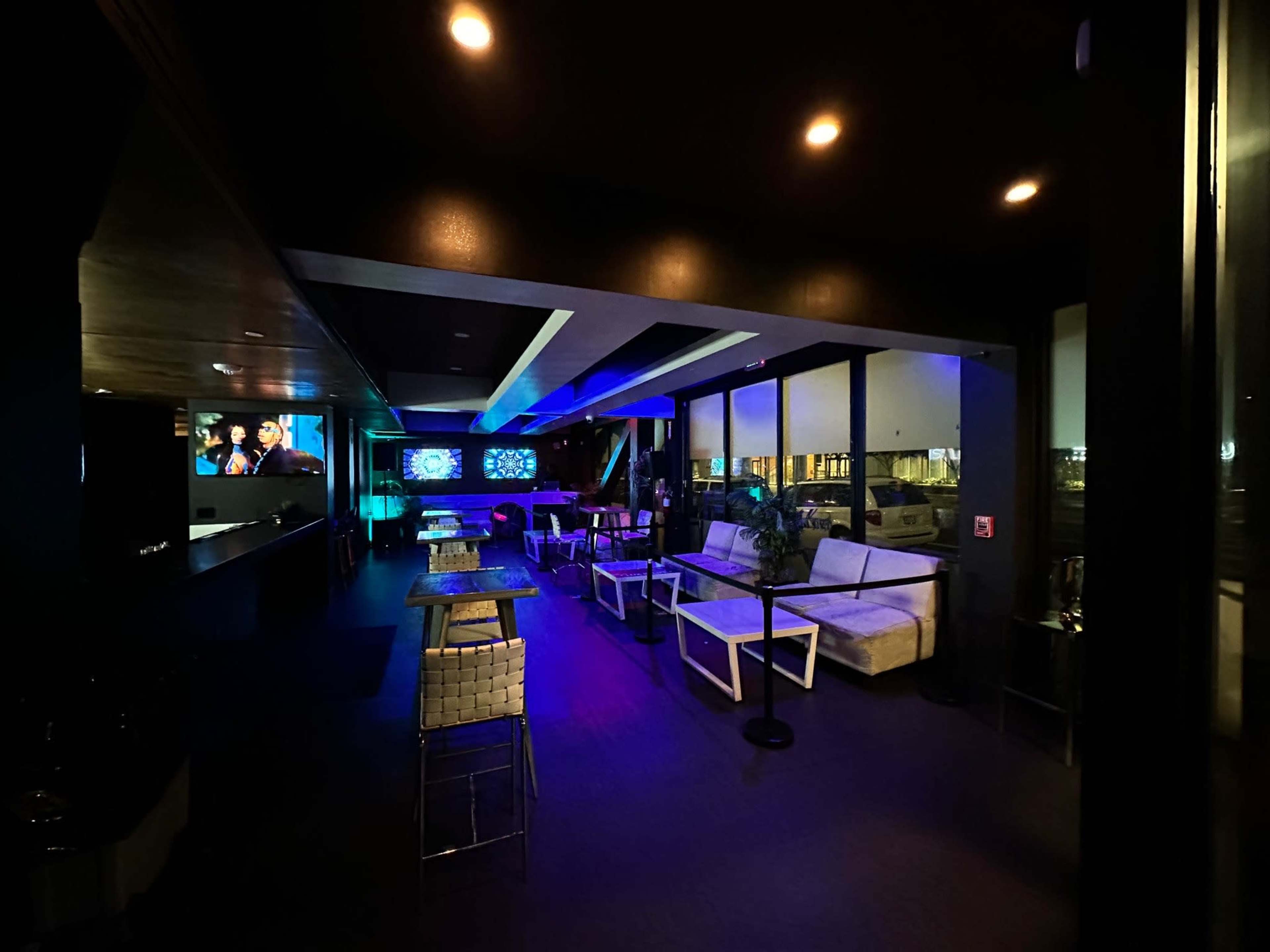 A modern lounge features comfortable seating, illuminated by colorful lights, with a bar area and screens displaying visuals.