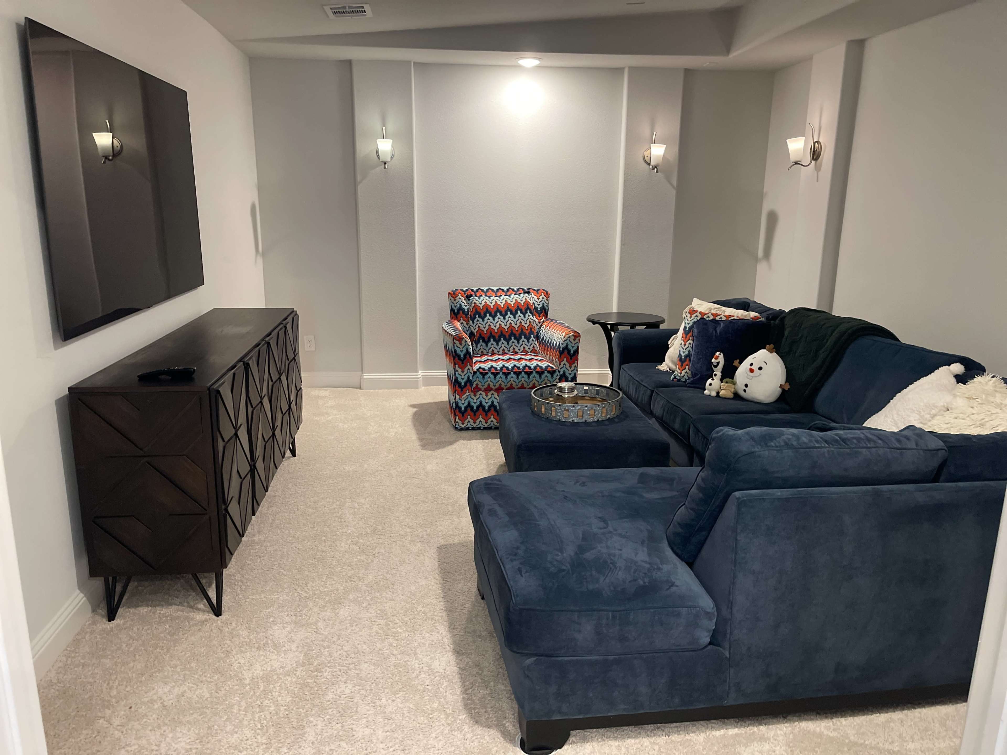 A cozy basement living room features a blue sectional sofa, a patterned chair, a wooden TV stand, and wall sconces, all set against light gray walls and plush carpet.