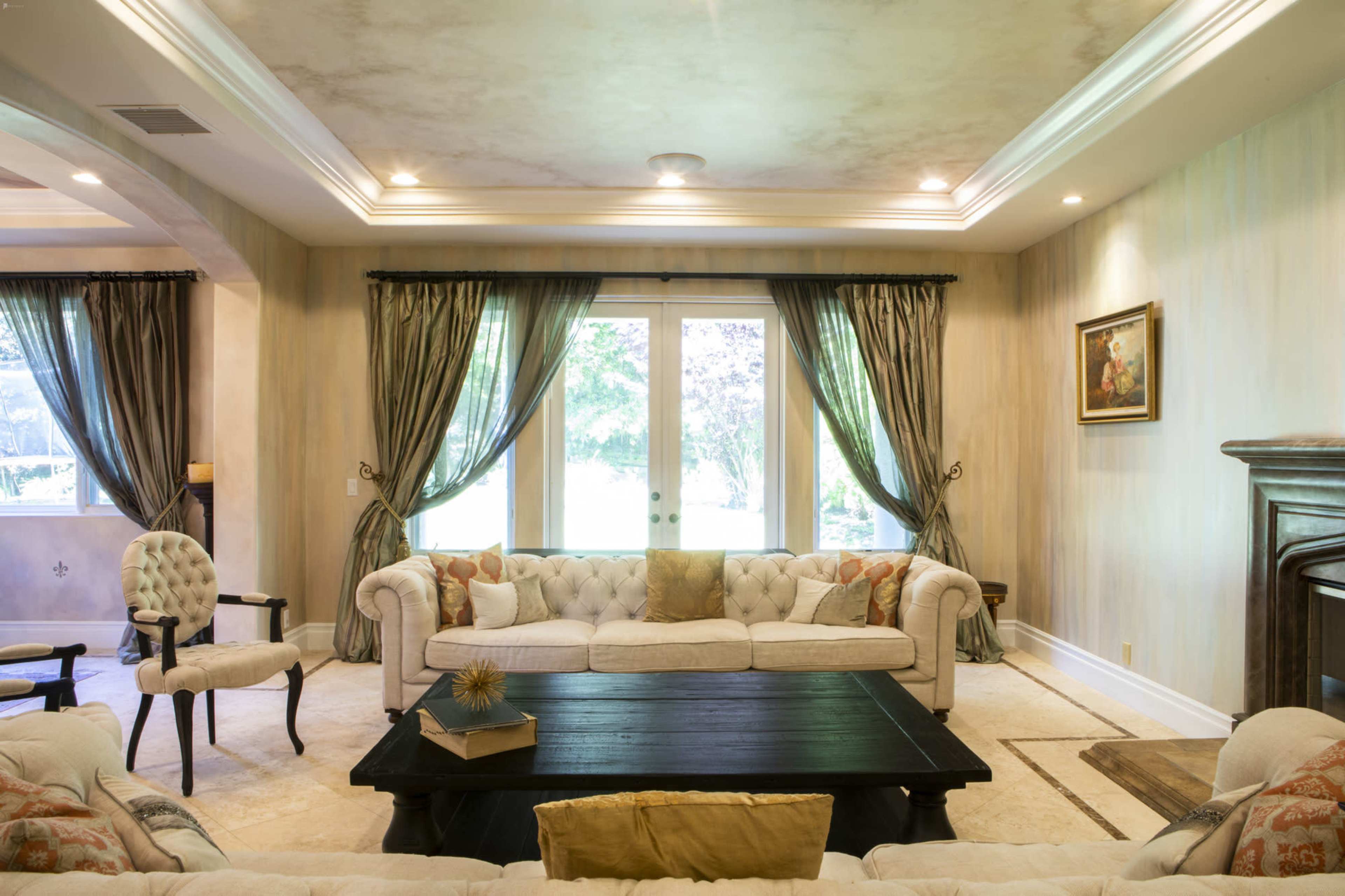A spacious living room features a set of beige tufted sofas, a dark wooden coffee table, and large windows draped with green curtains.
