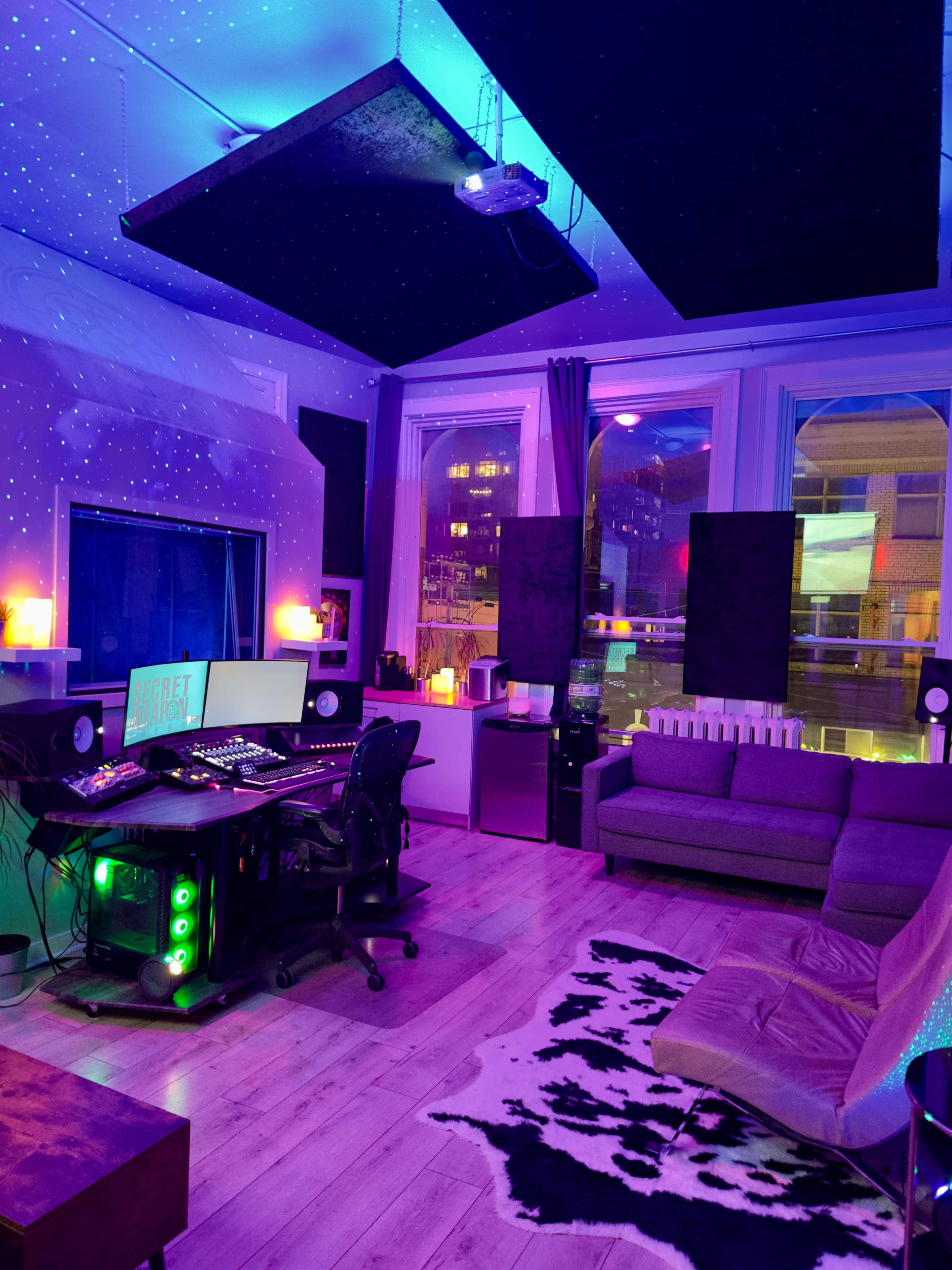 A modern studio space with a computer desk, a couch, and ambient lighting, featuring a projected starry ceiling and sound panels on the walls.