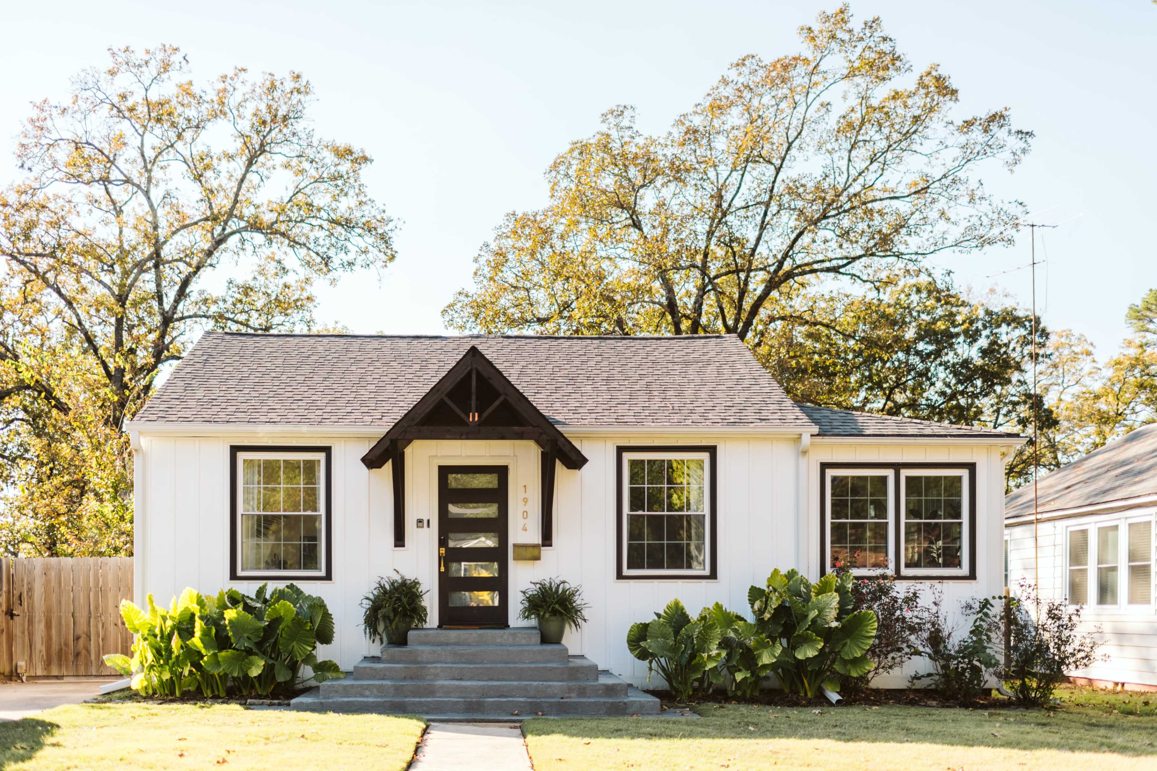 A single-story white house with a black-trimmed front door and large windows is surrounded by green plants and trees.