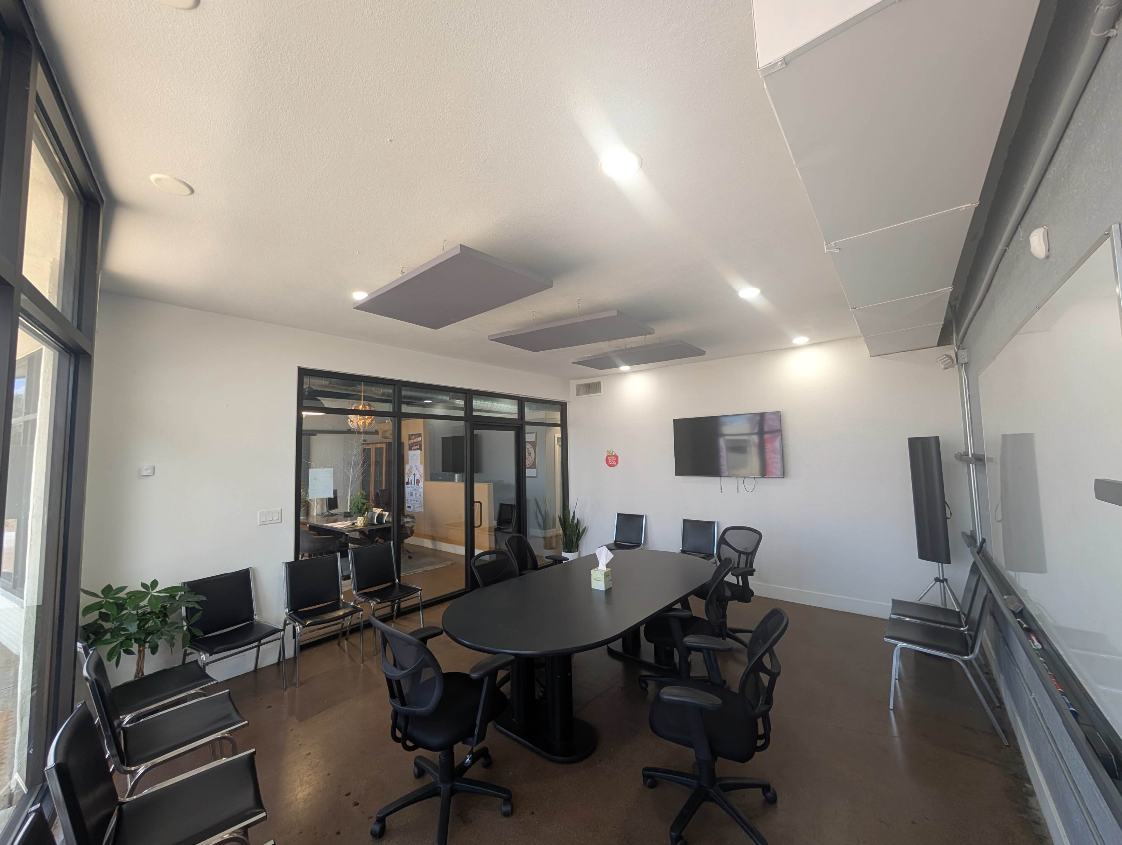 The image shows a modern conference room with a large black oval table surrounded by ergonomic chairs and a television screen mounted on the wall.