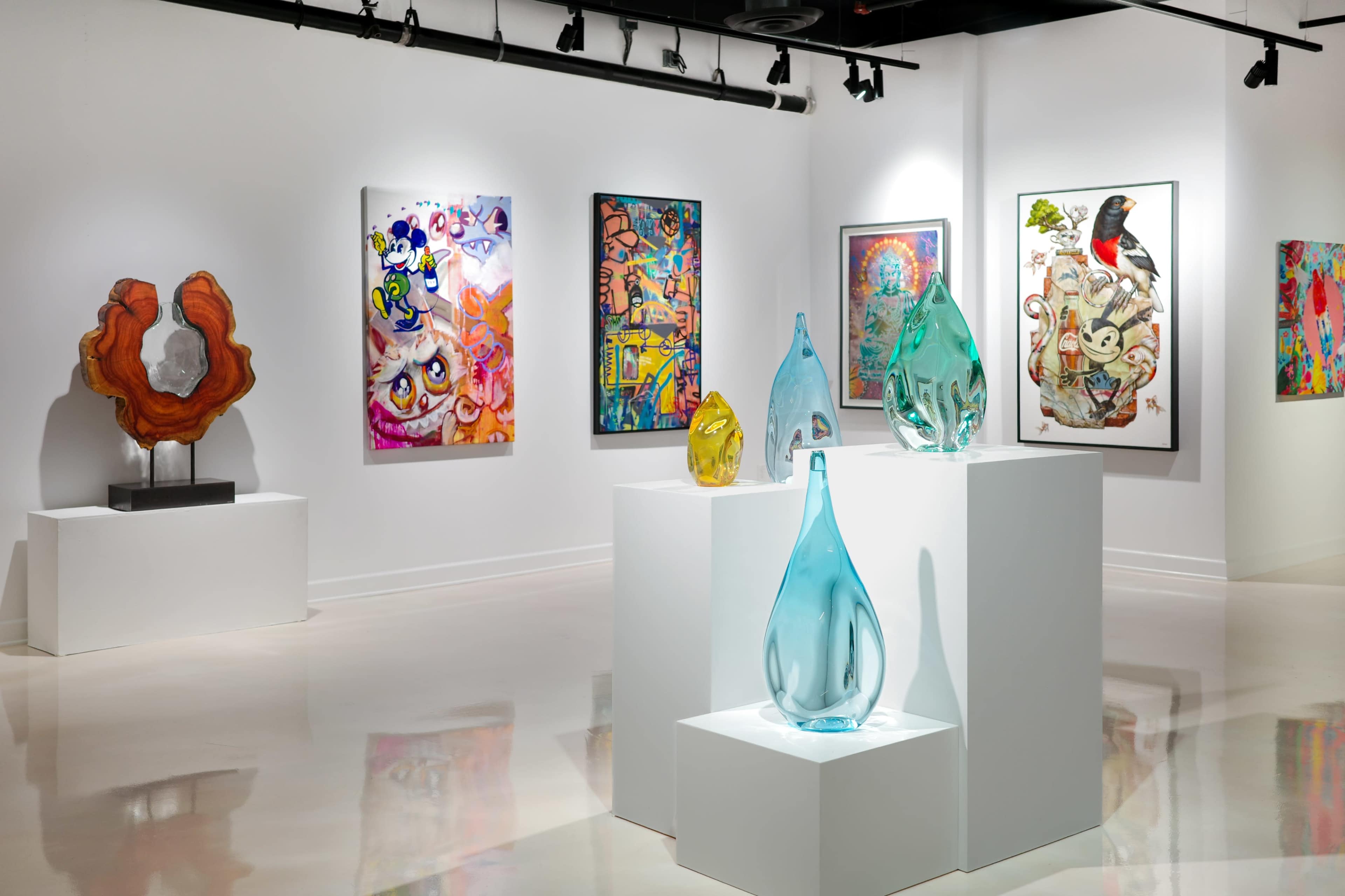 The image shows an art gallery with colorful paintings and glass sculptures displayed on white pedestals.
