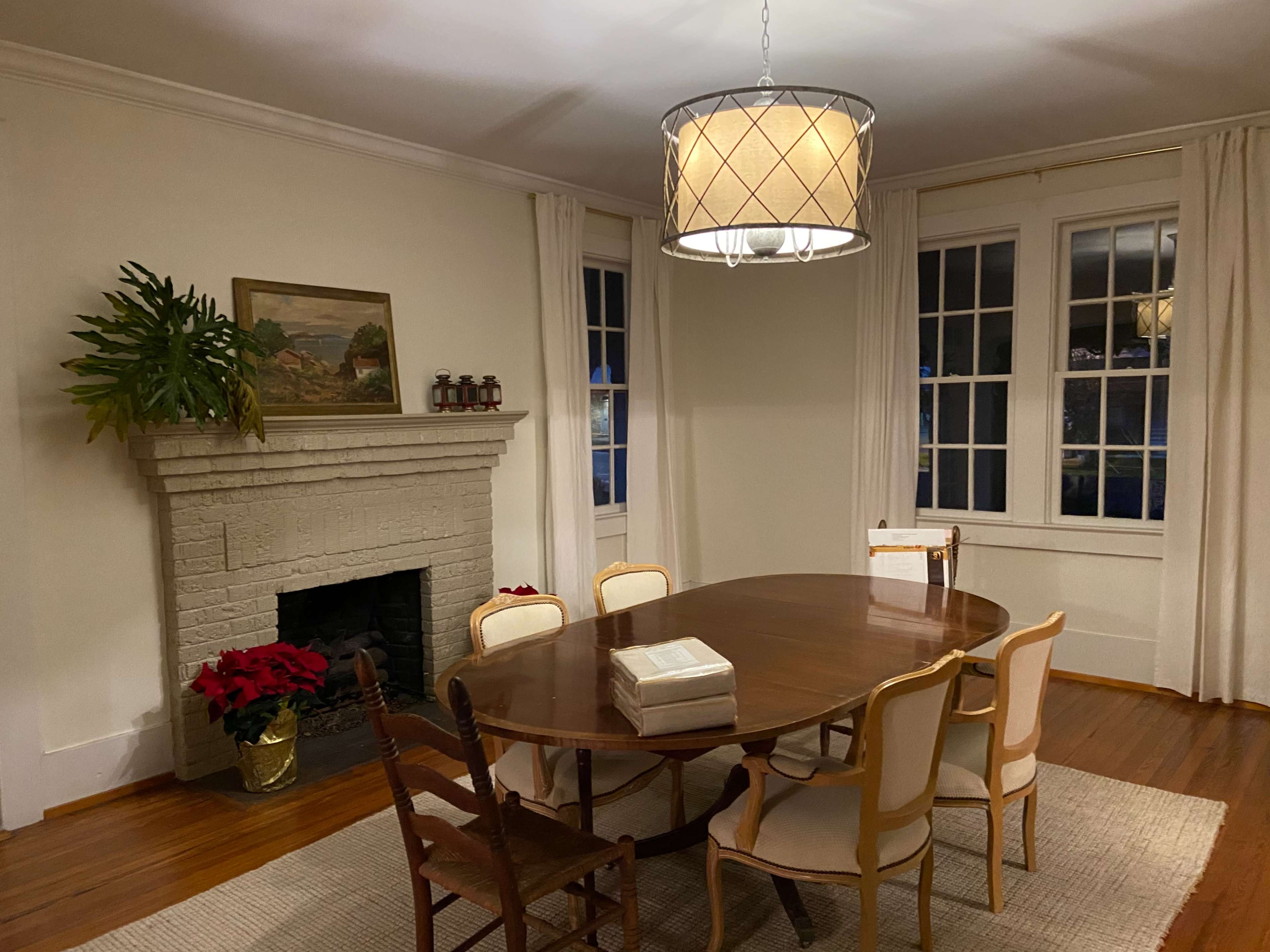 A dining room with a round table surrounded by six chairs, a fireplace with a painting above it, and two large windows with white curtains.