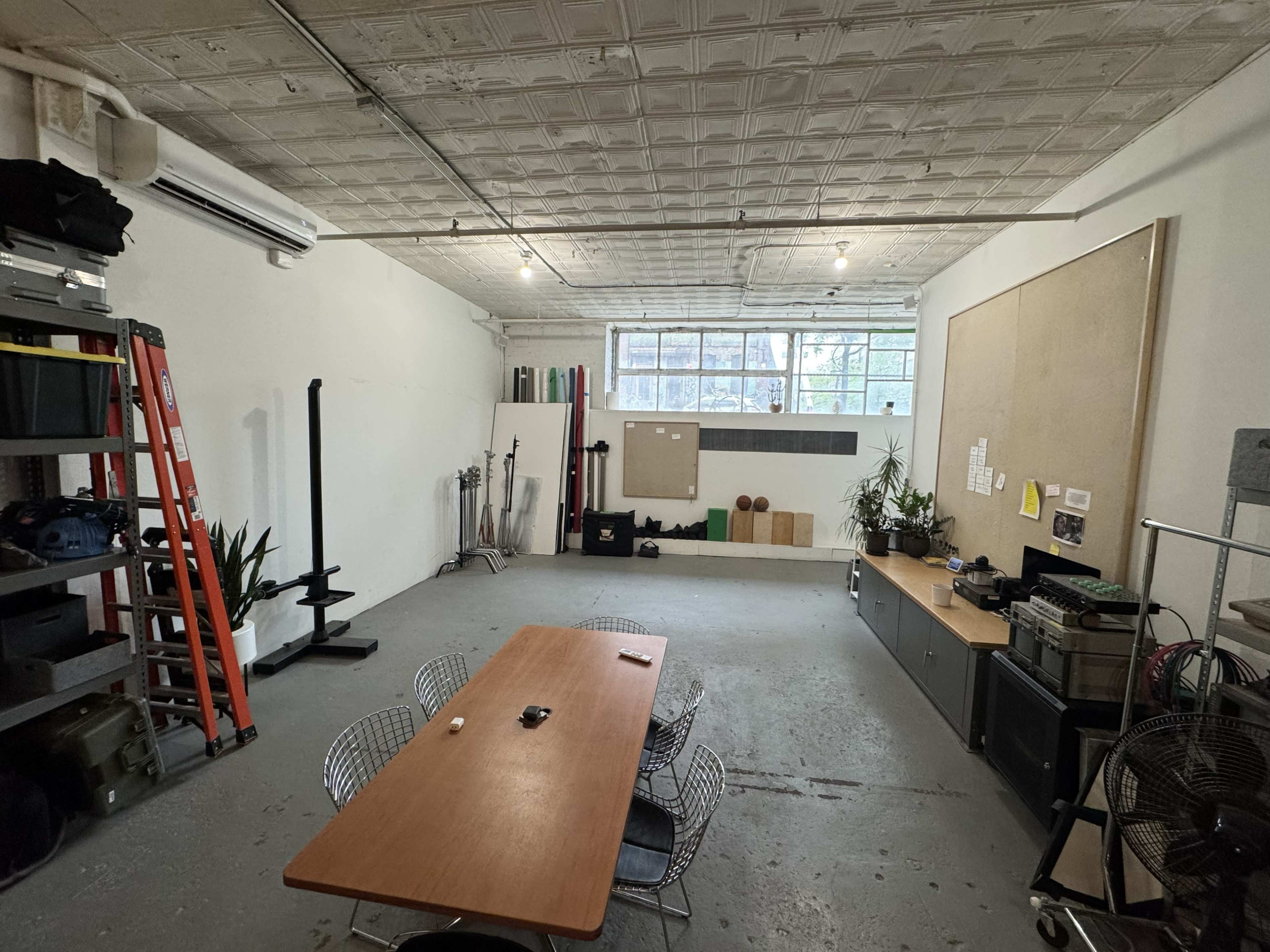 A spacious, well-lit room features a table with chairs, storage shelves, a ladder, and various equipment along the walls.