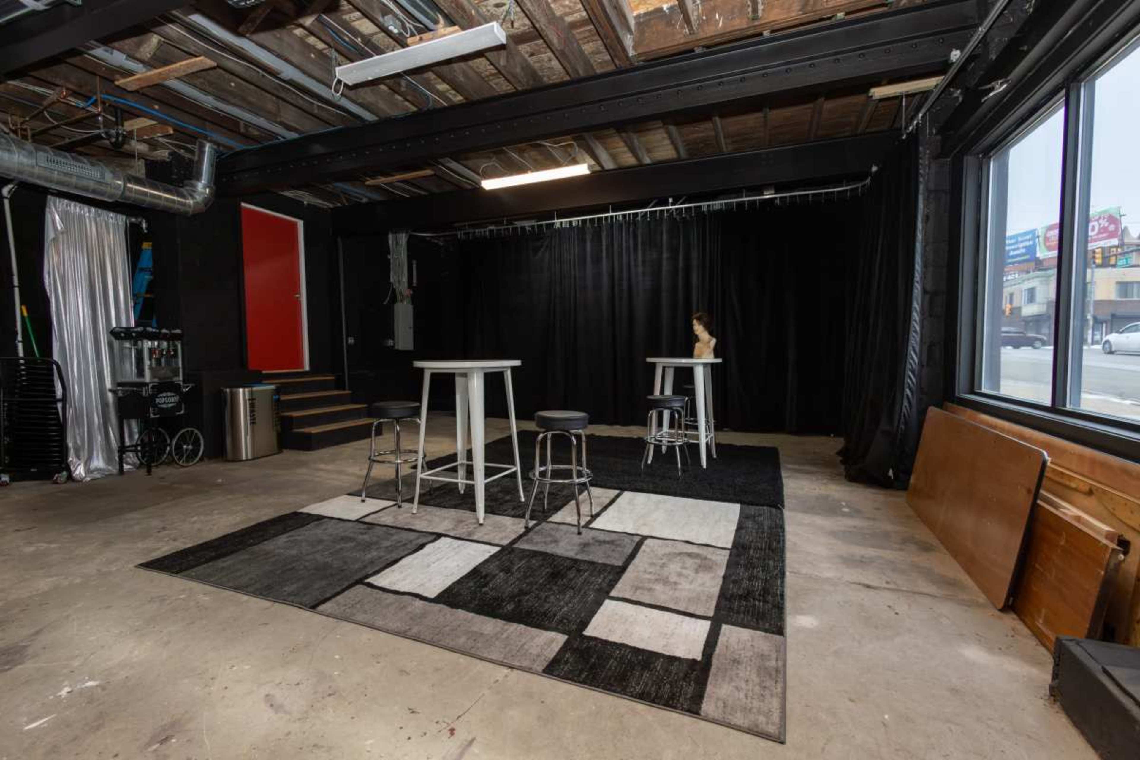The image shows an empty studio space with a concrete floor, two tall white tables surrounded by black and gray patterned chairs, and black curtains in the background.