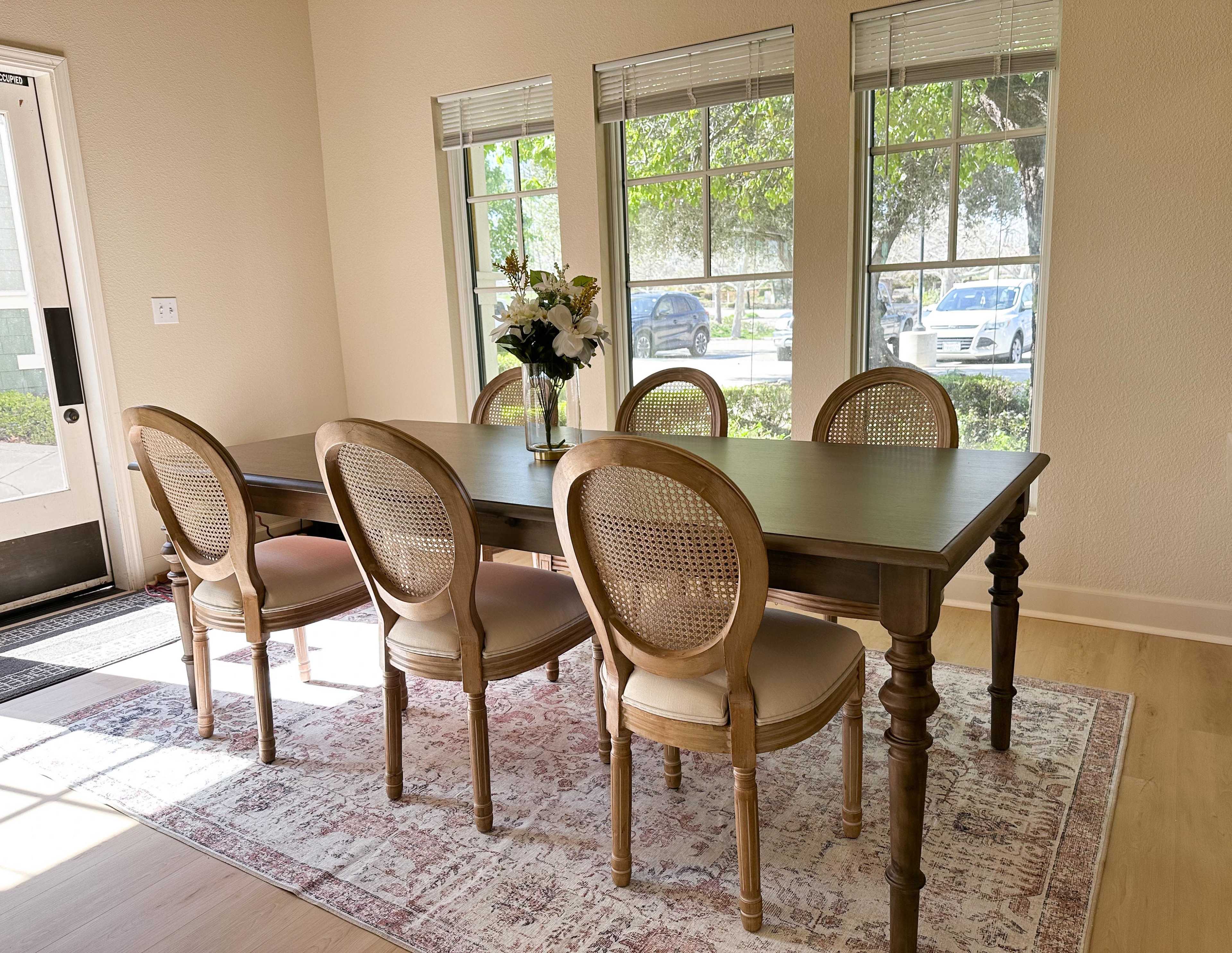 A rectangular dining table with six cane-backed chairs is positioned under large windows in a brightly lit room.