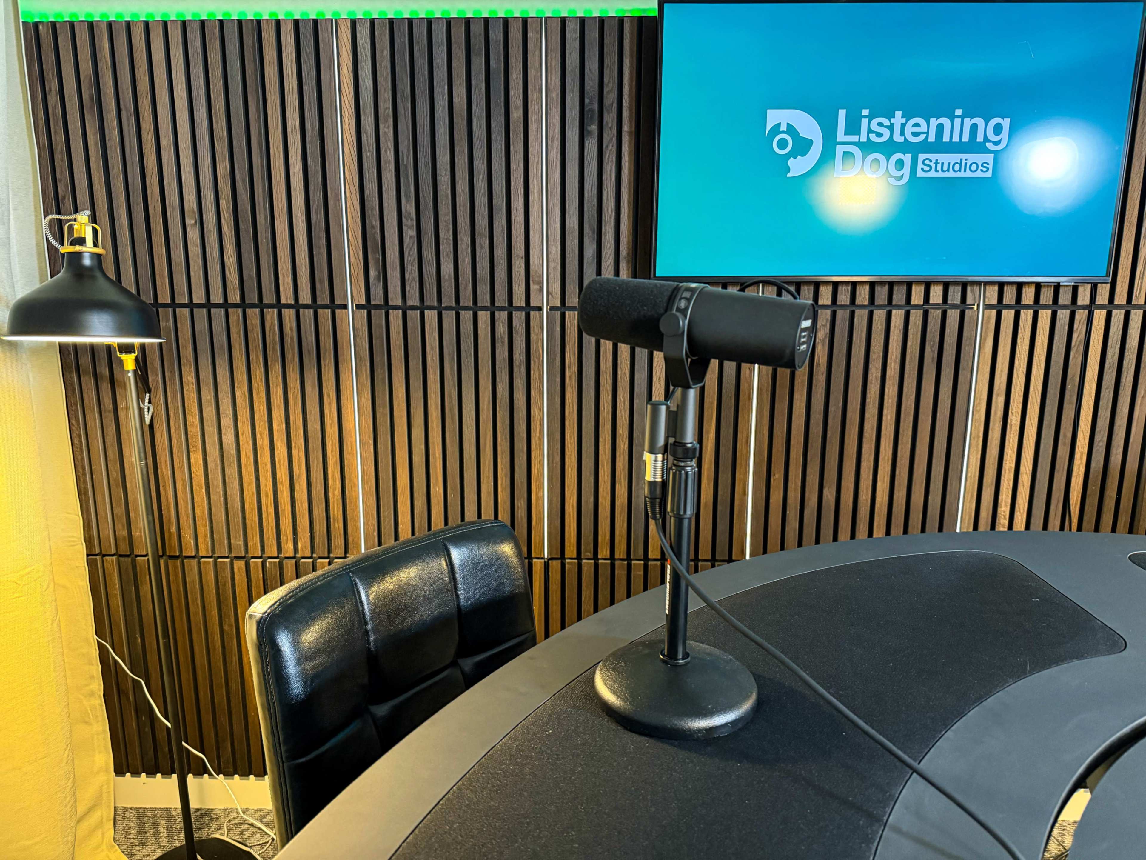 The image shows a microphone on a stand positioned on a round table, with a black chair nearby and a screen displaying "Listening Dog Studios" in the background.
