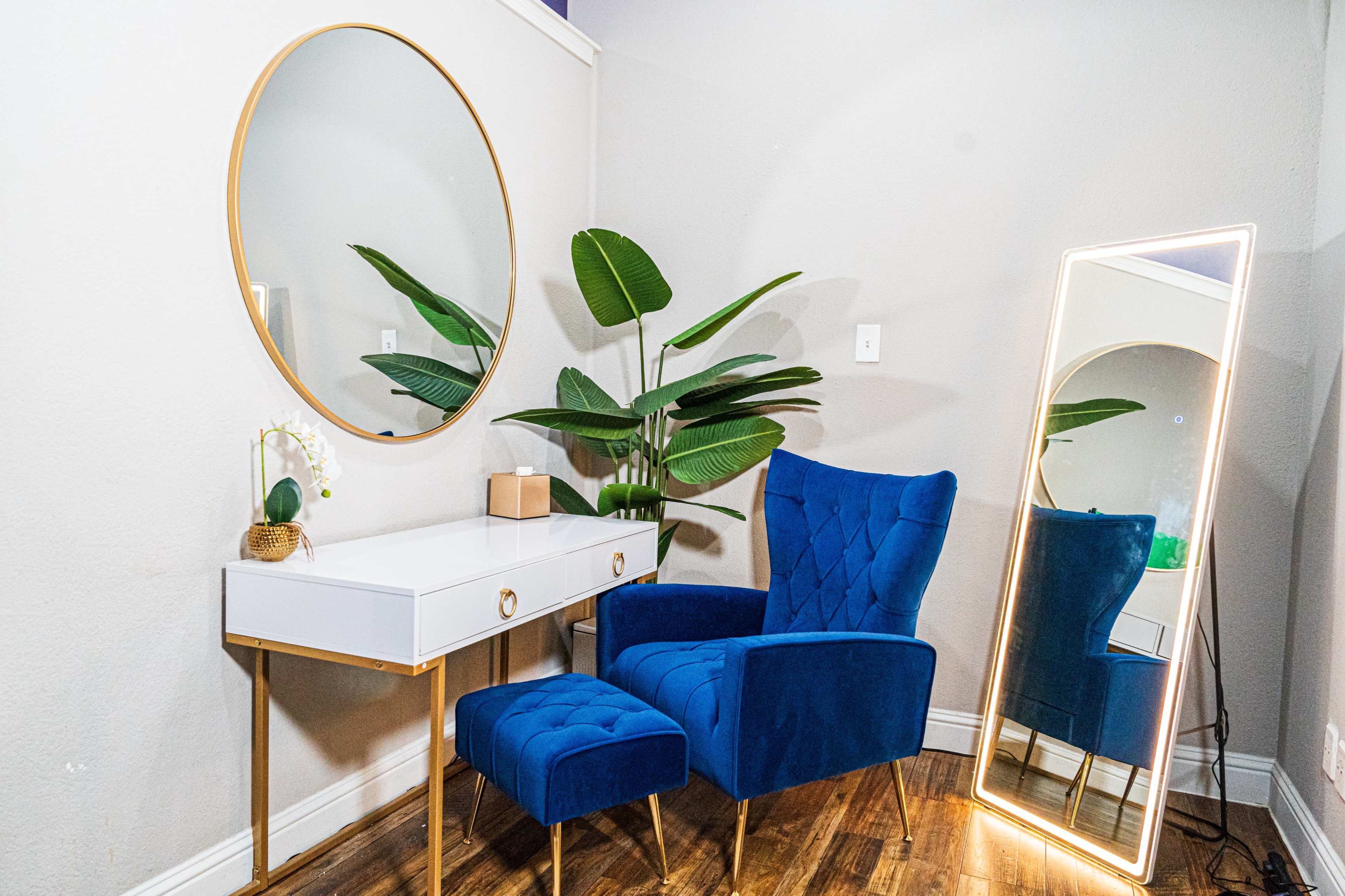The image shows a modern corner featuring a blue chair and ottoman next to a white vanity with a round mirror and a tall mirror, accompanied by a green plant.