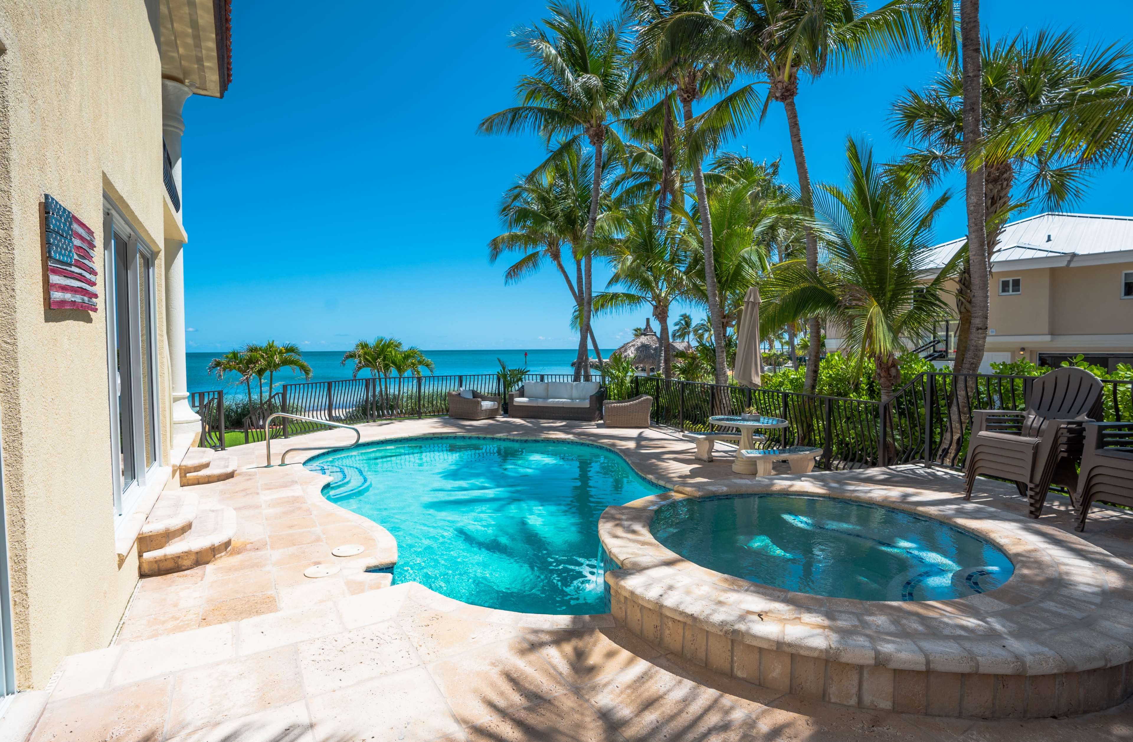 A backyard features a swimming pool and hot tub surrounded by palm trees, overlooking the ocean.