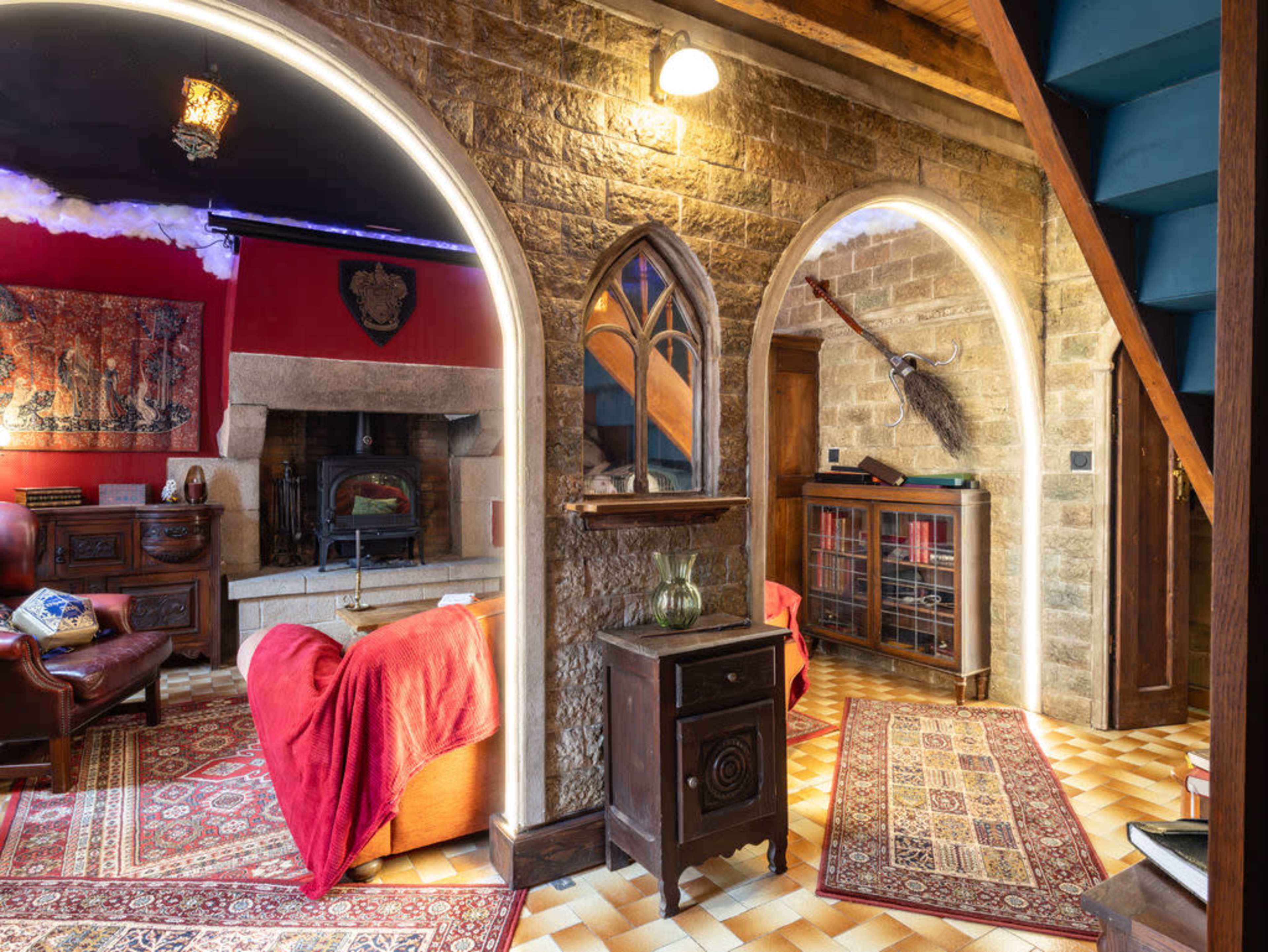 The image shows a cozy interior with stone walls, an arched doorway, a fireplace, a red couch, and decorative rugs.