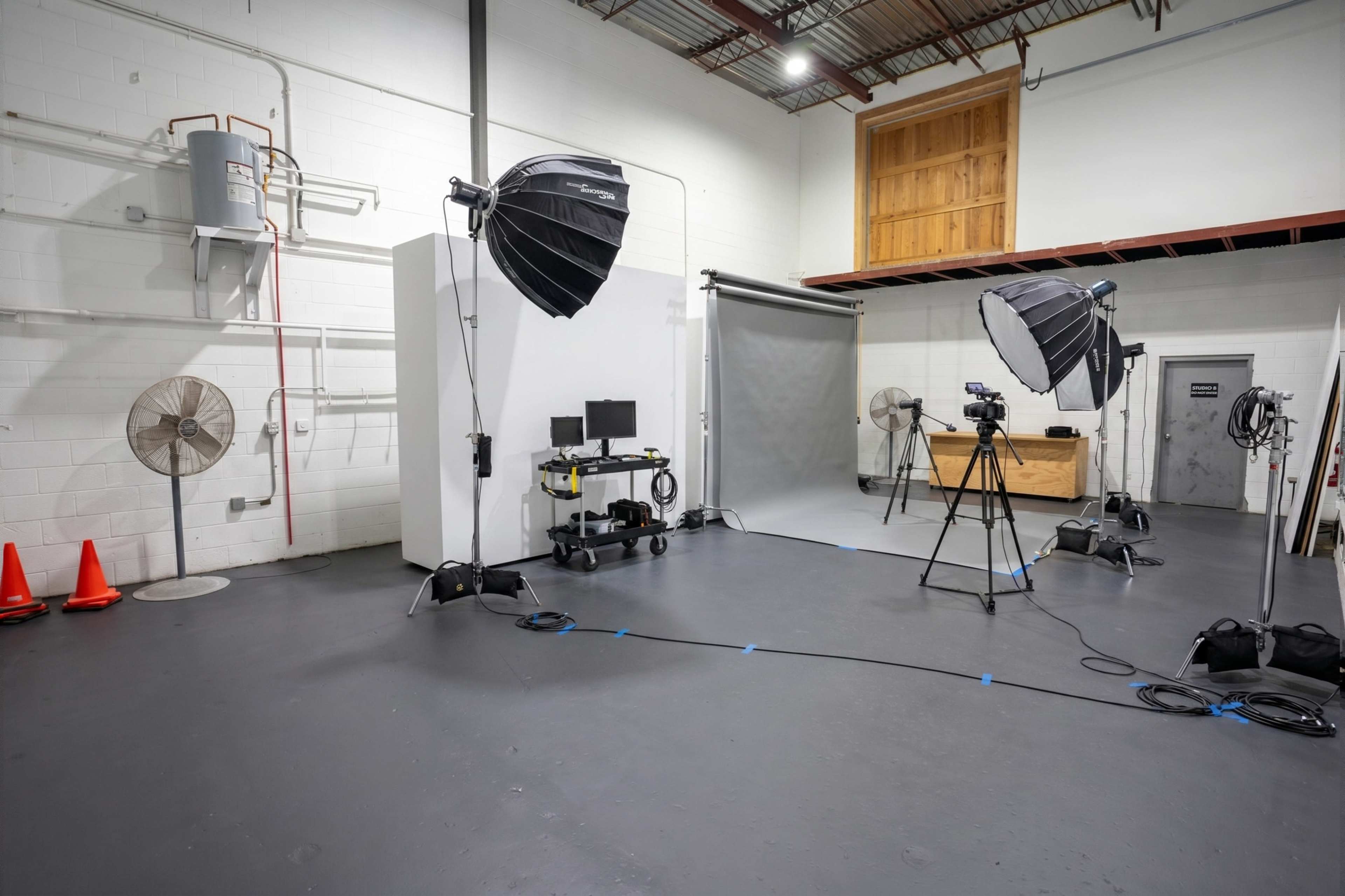 The image shows a spacious photography studio with lighting equipment, a camera setup, a backdrop, and a few fans arranged around the room.