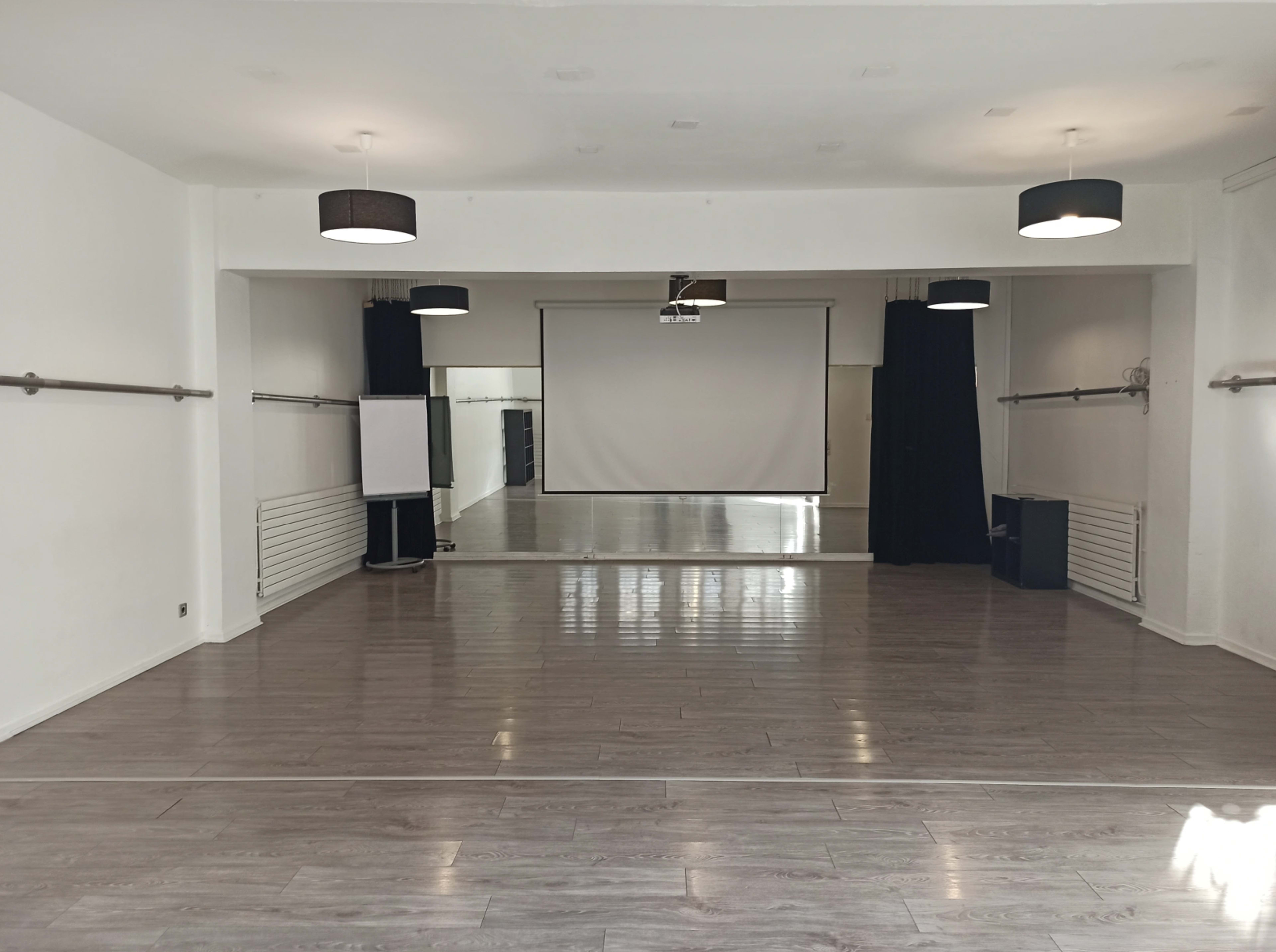 A spacious, empty room with wooden flooring, wall-mounted ballet barres, a projector screen, and multiple light fixtures.